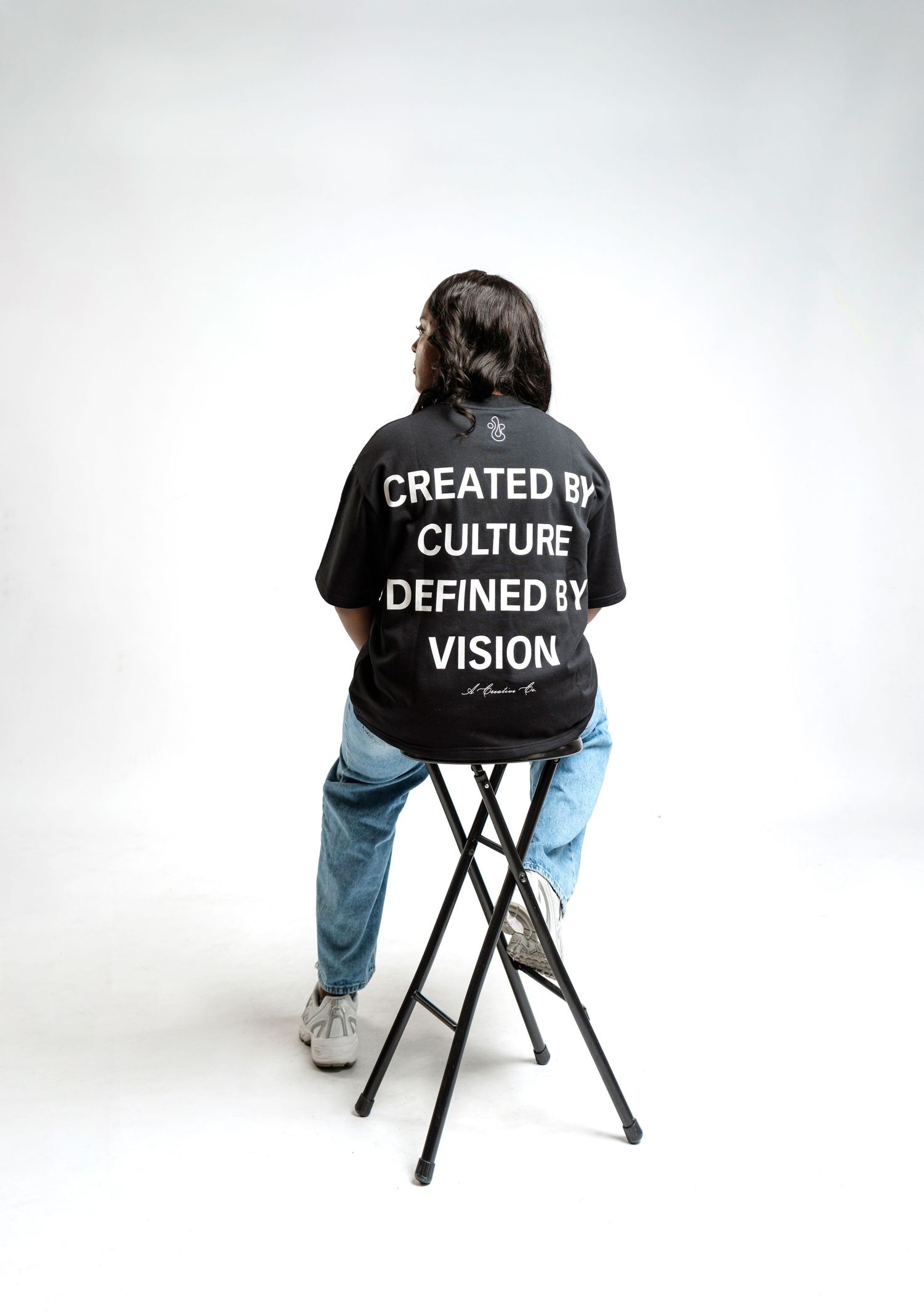 Person wearing black t-shirt with white text sits on stool against white backdrop, showing back design.