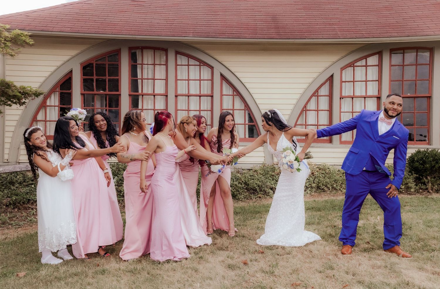 Wedding party in pink dresses and blue suits posing playfully outside building.