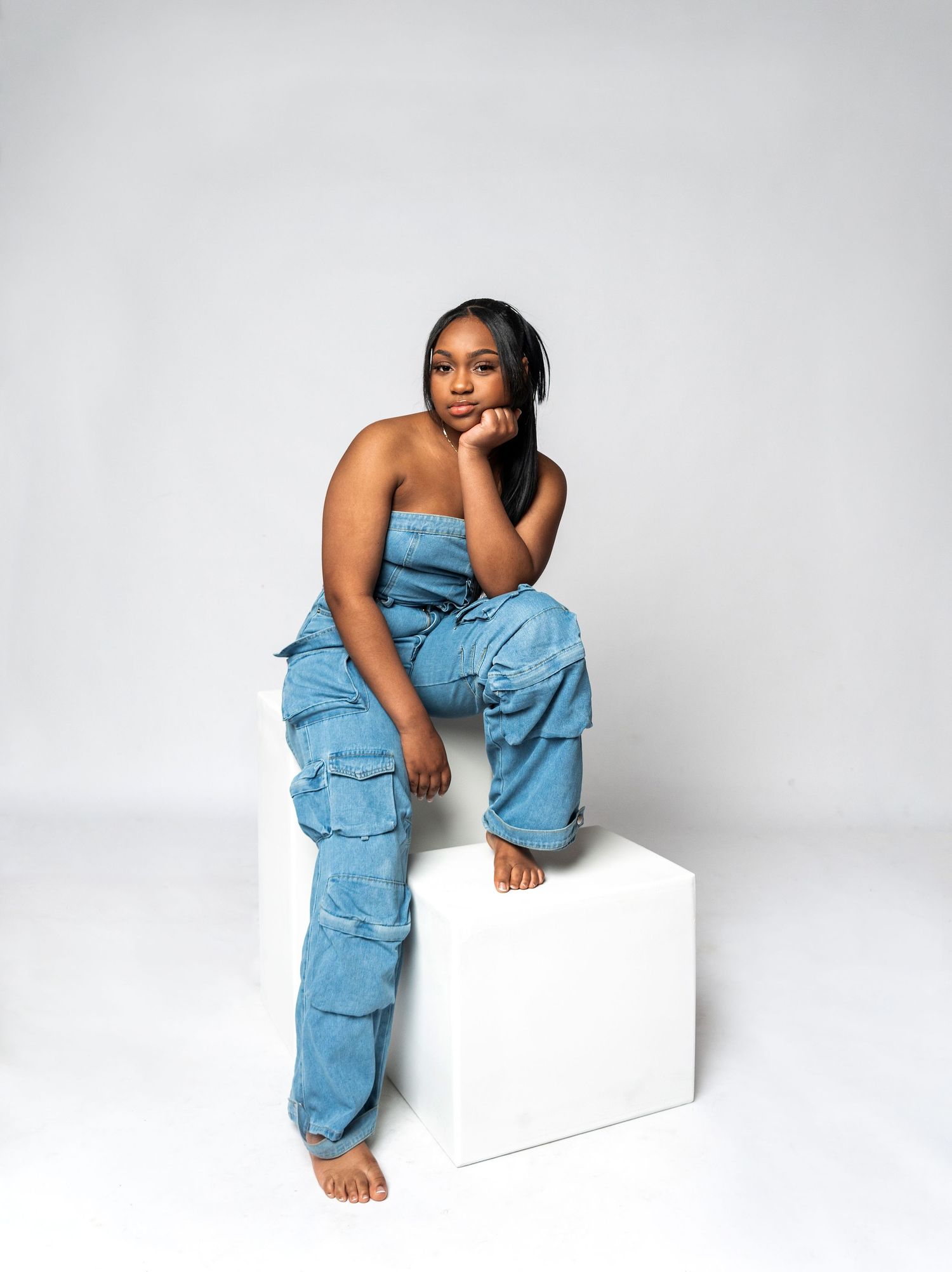 Model poses on white backdrop wearing blue denim strapless jumpsuit in fashion editorial series.