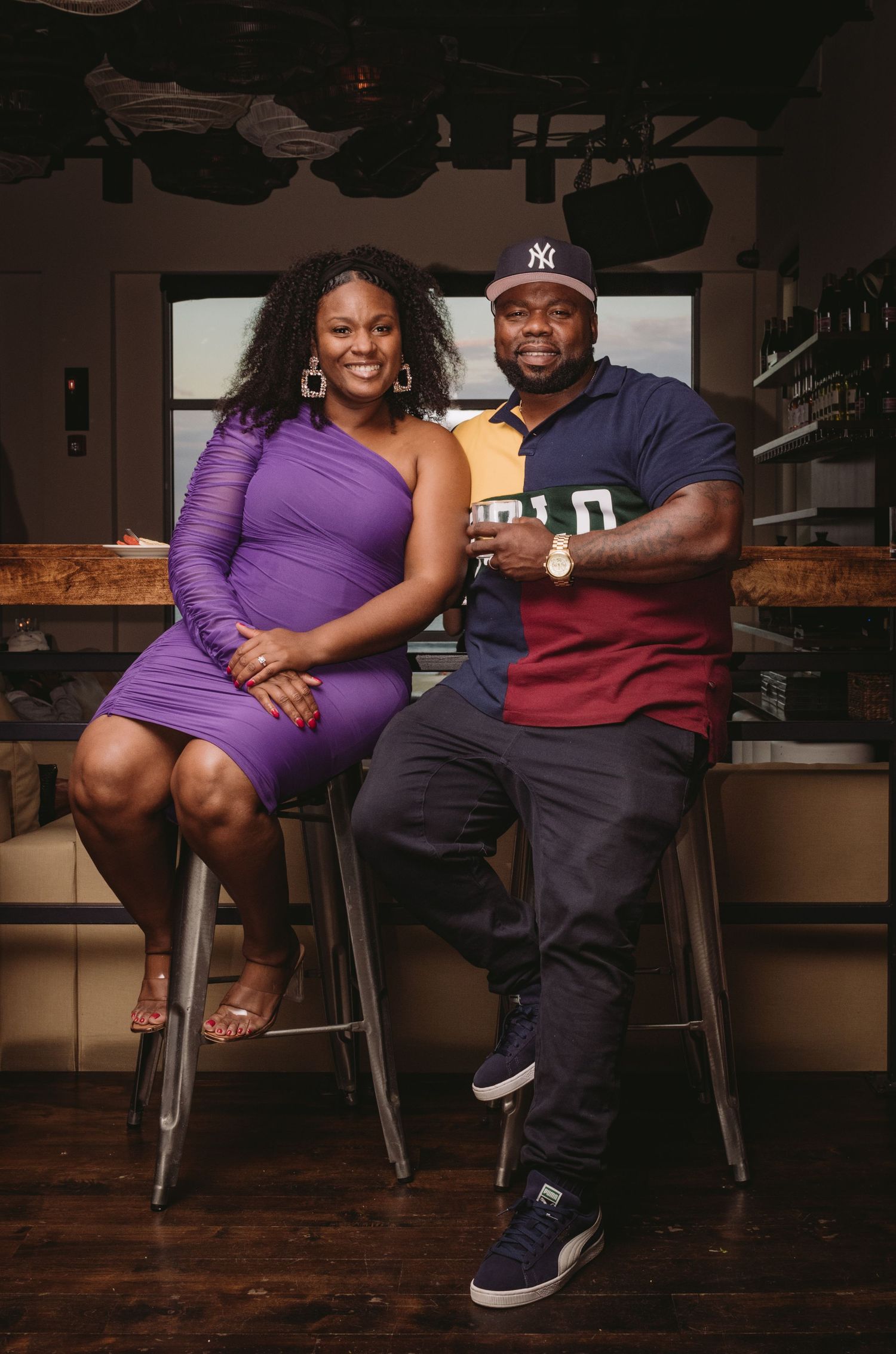 A couple wearing purple and black sits together on bar stools in a dimly lit room with wooden flooring.