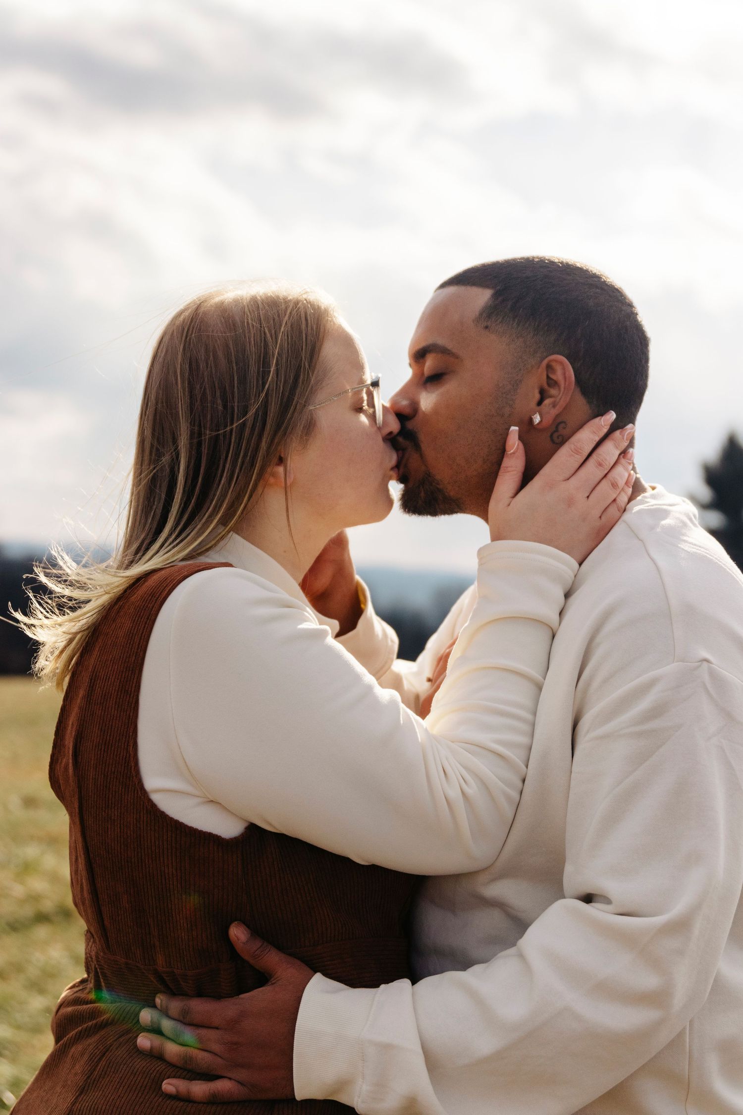 A romantic couple share intimate kisses while wearing cream colored sweaters against a cloudy sky backdrop.