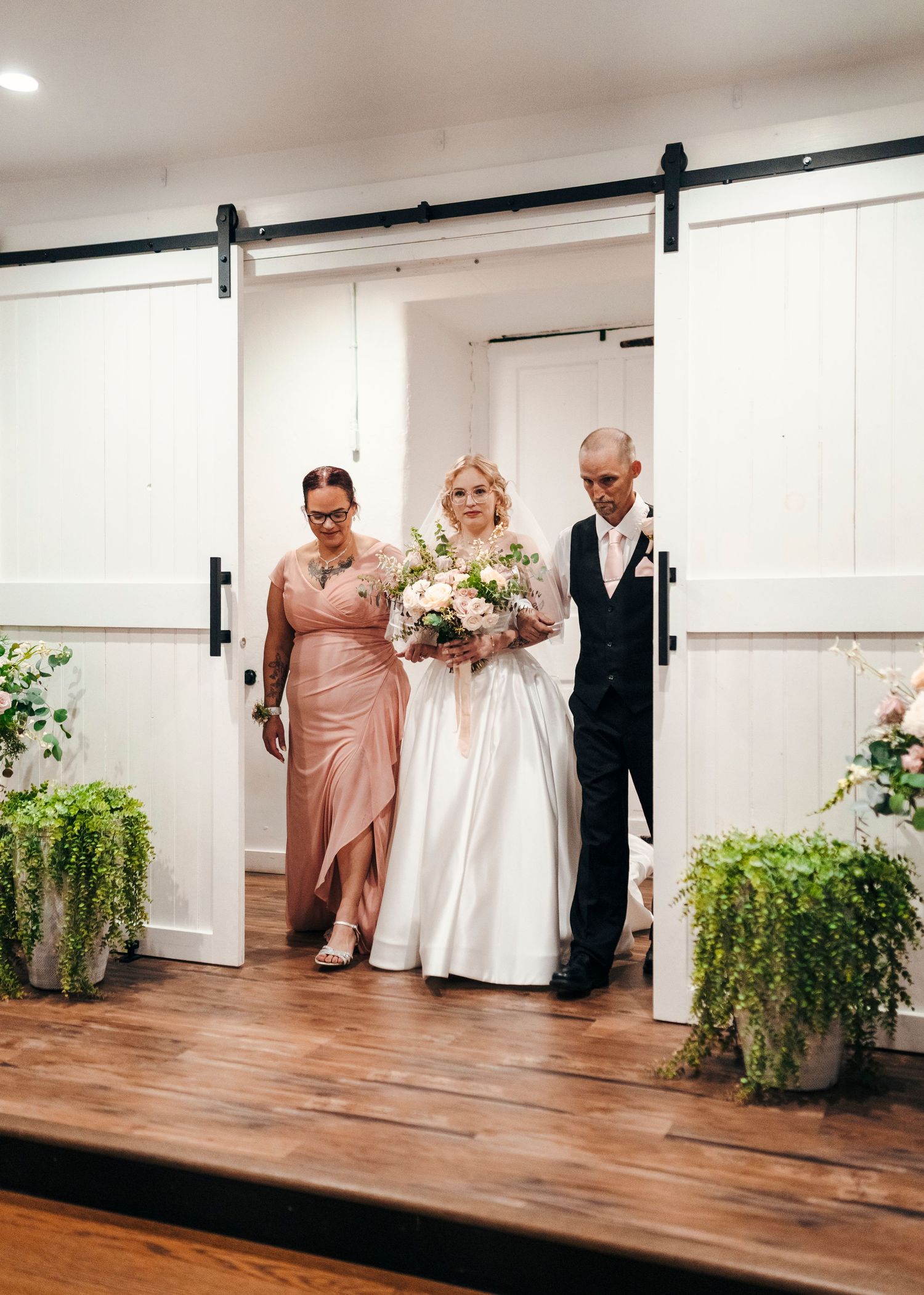 Bride in white gown holds bouquet flanked by two escorts through white barn doors at rustic wedding venue.