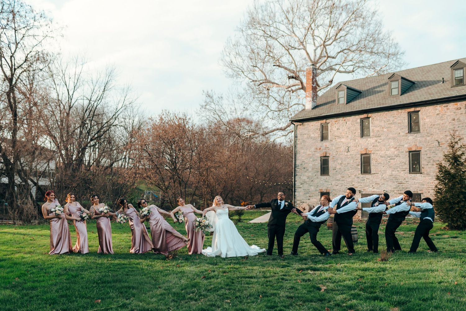 Fun wedding party photo with bridesmaids pulling bride and groom away from groomsmen outdoors.