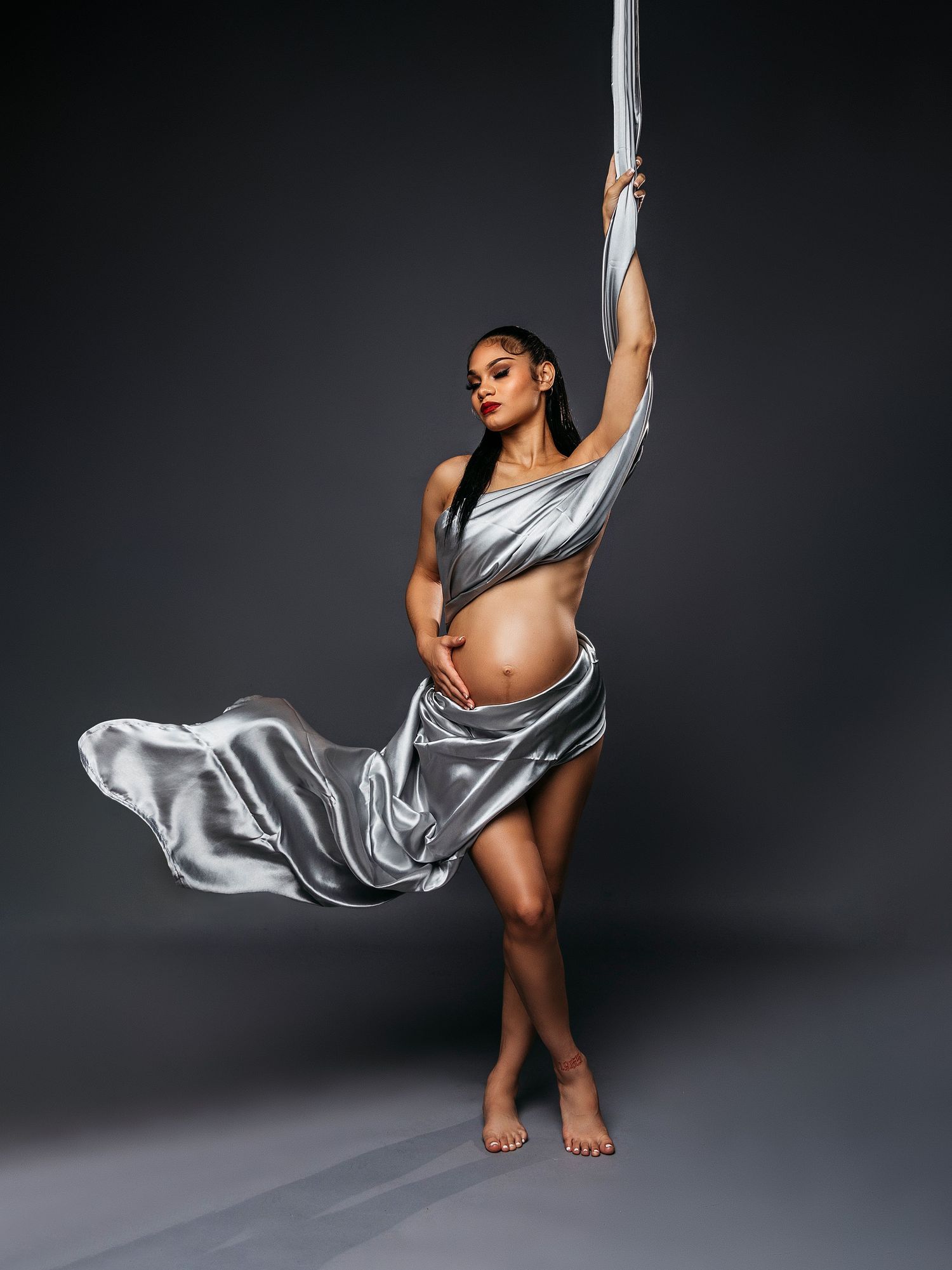 Pregnant dancer in flowing silver fabric performs graceful pose against dark background.
