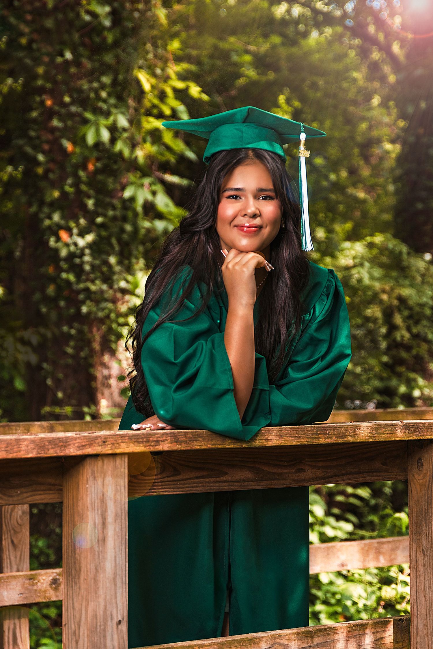 Outdoor graduation portrait of student in emerald green cap and gown leaning on wooden railing.