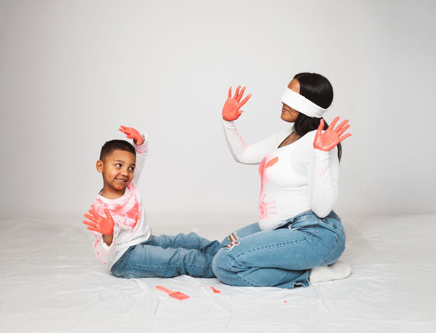 A woman and child sit together covered in pink paint, both smiling with hands raised in a playful photo shoot.