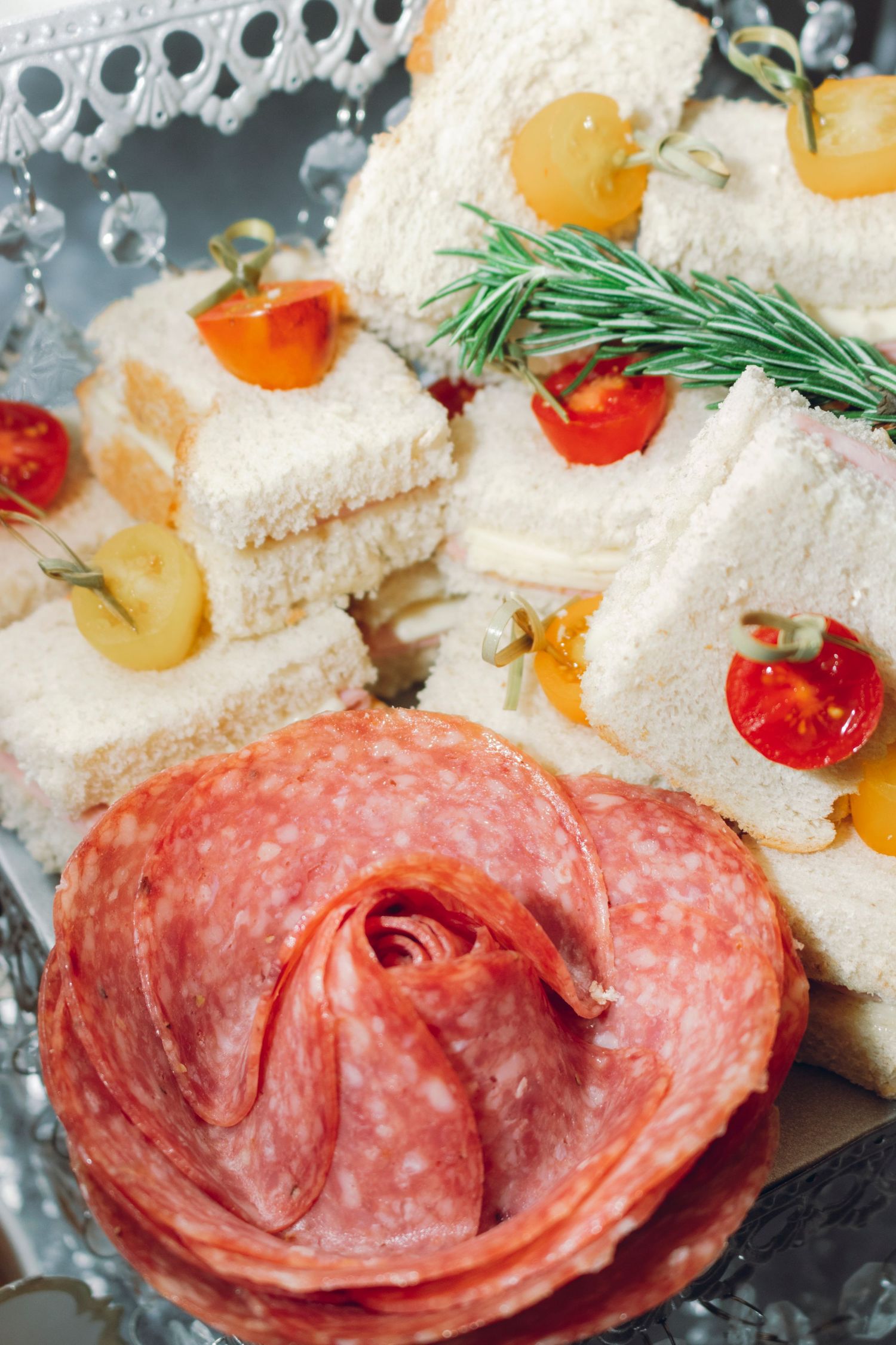 Close-up of thinly sliced salami arranged in an overlapping pattern with finger sandwiches and garnishes in the background.