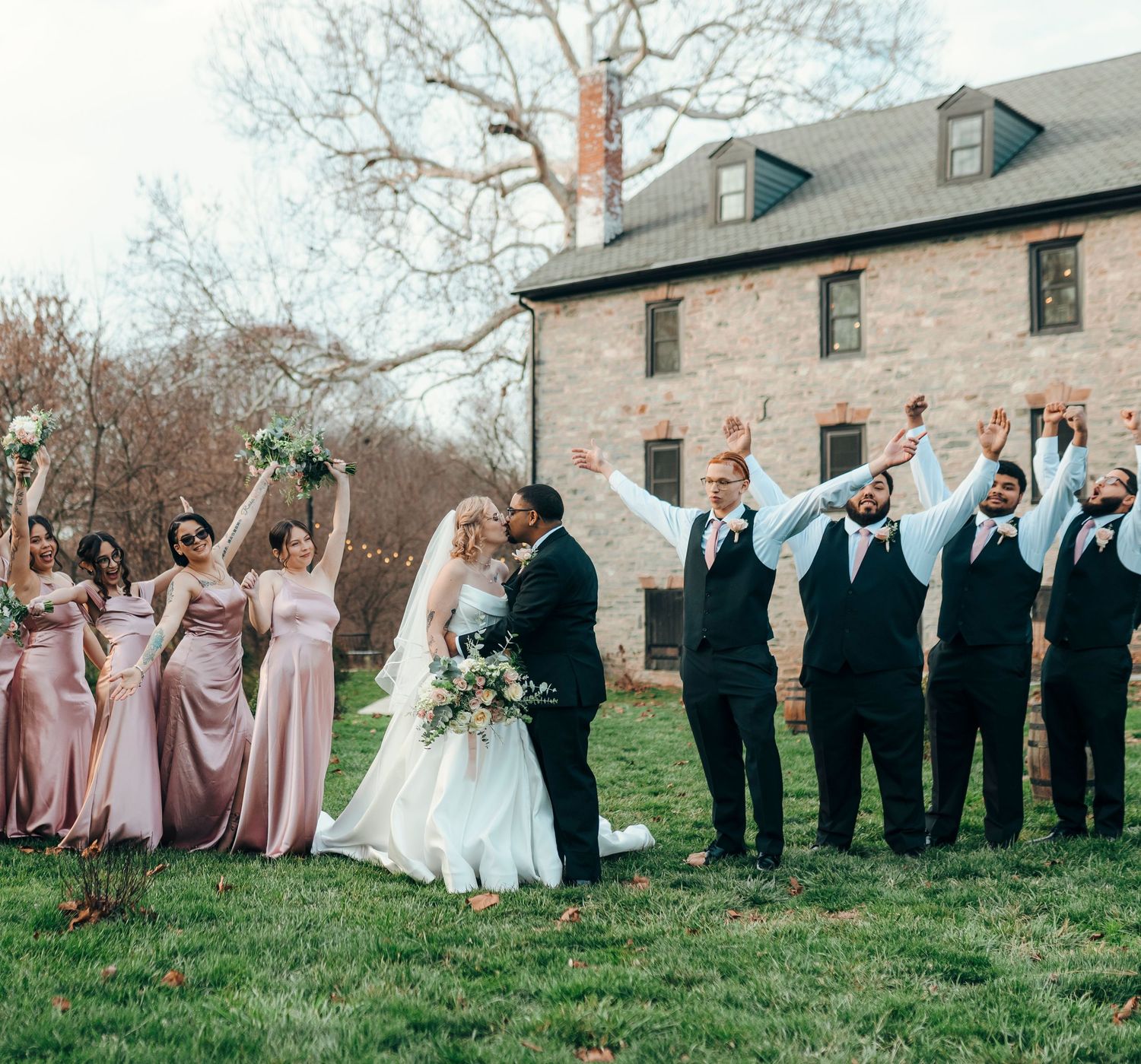 Wedding party celebrates outdoors, bridesmaids in pink gowns and groomsmen in vests cheer as couple kisses near stone building.
