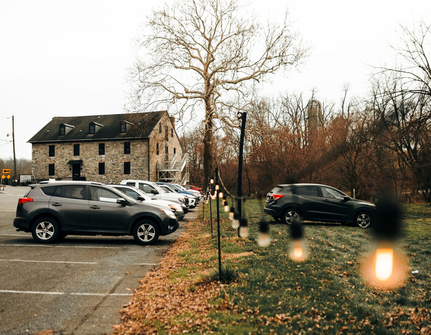 Cars parked in a lot beside a historic stone building with string lights and bare trees on a cloudy day.