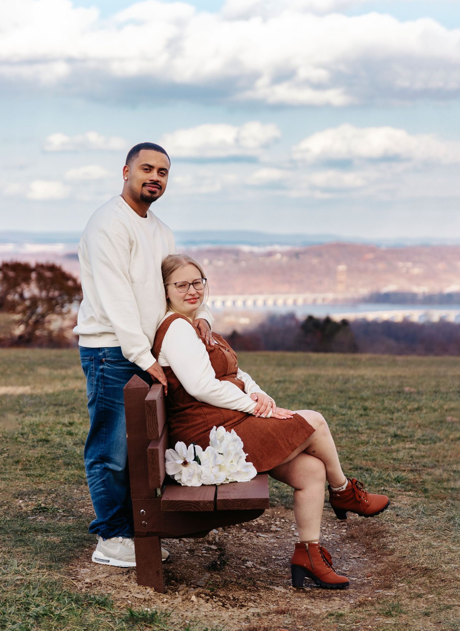 A couple shares a sweet moment together sitting on a vintage brown leather chair in an open grassy field at sunset.