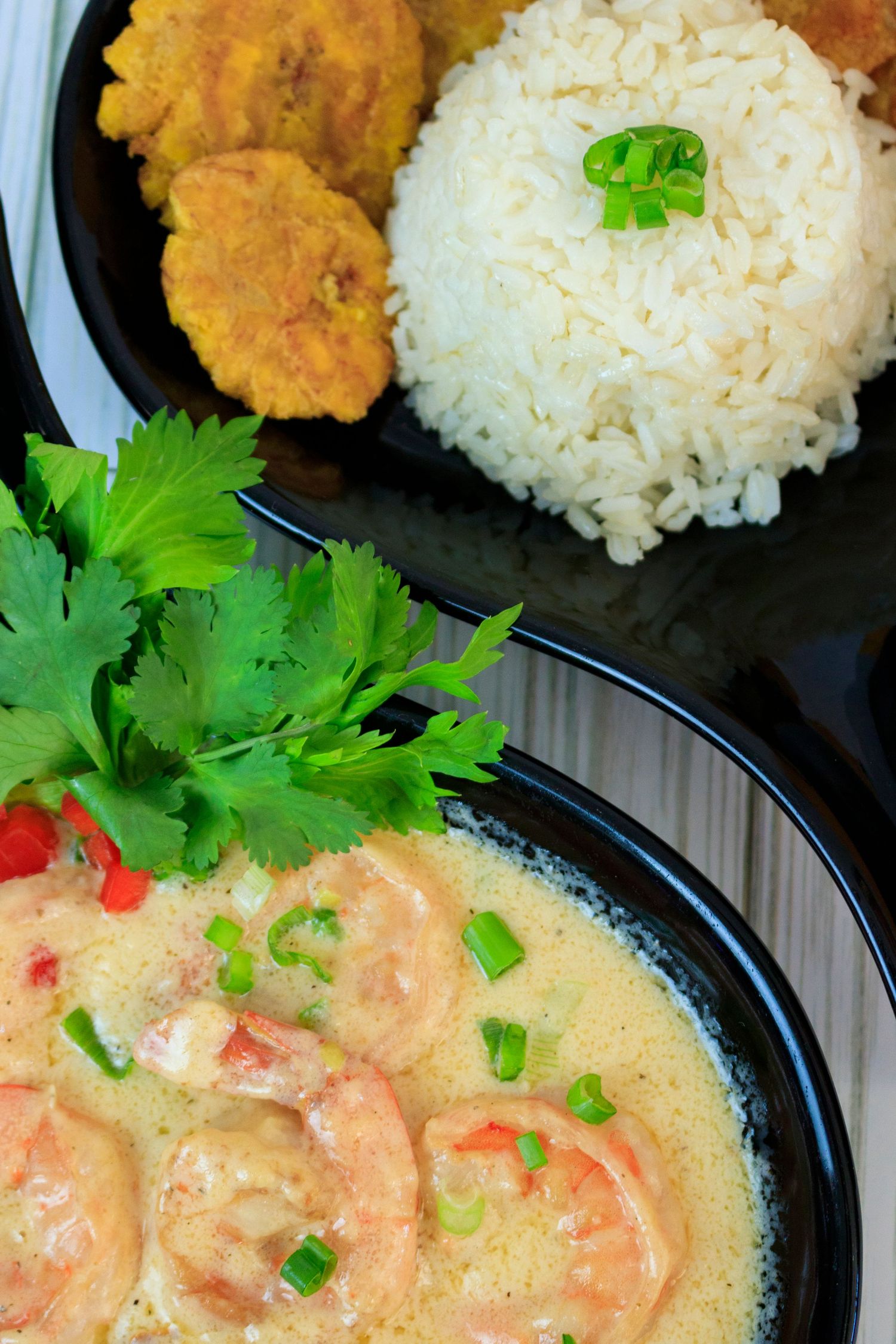 Creamy shrimp curry with white rice and tostones garnished with cilantro and green onions on a black plate.