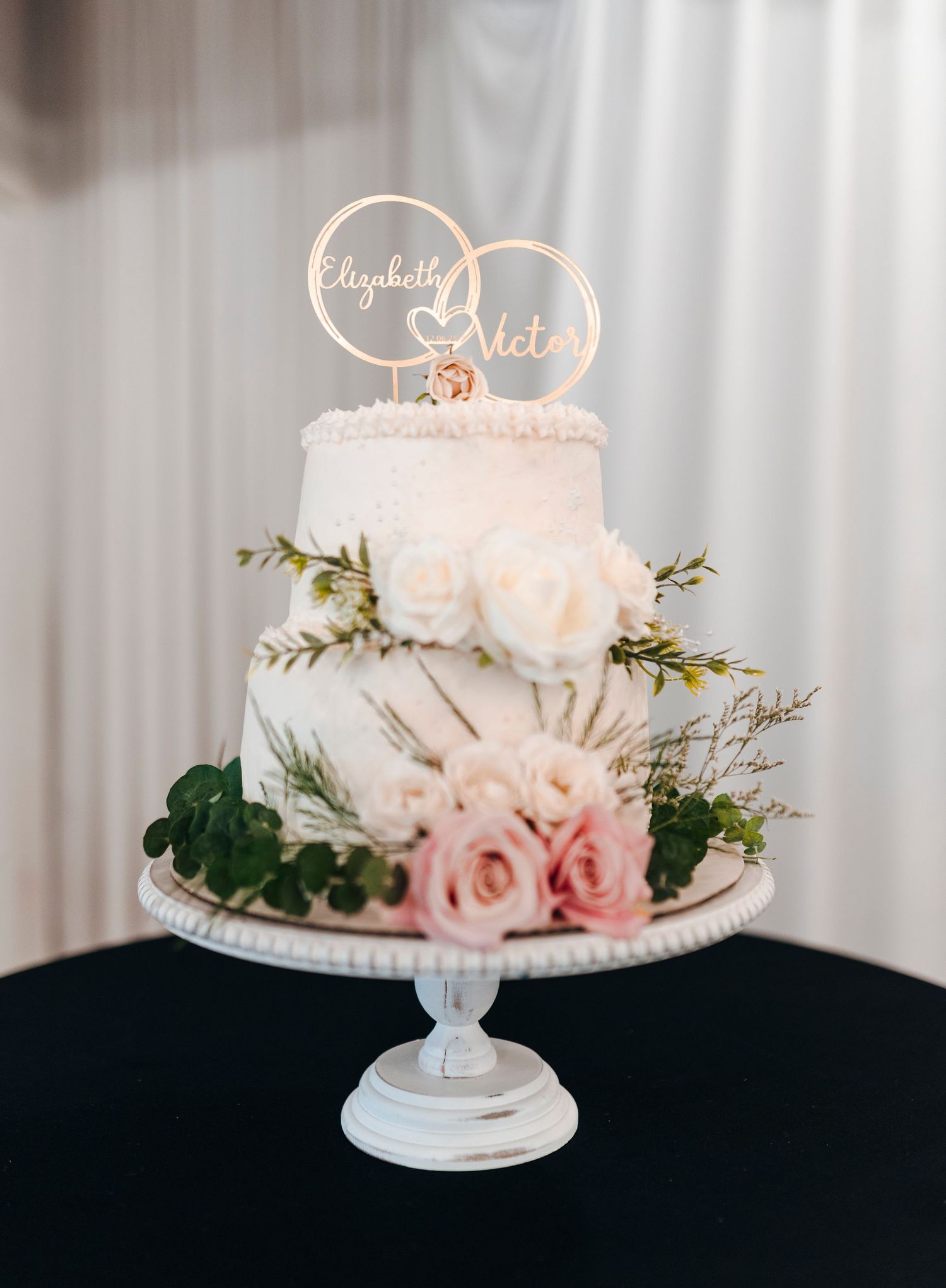 Elegant two-tier white wedding cake with blush roses, greenery, and gold personalized topper reading Elizabeth and Victor.