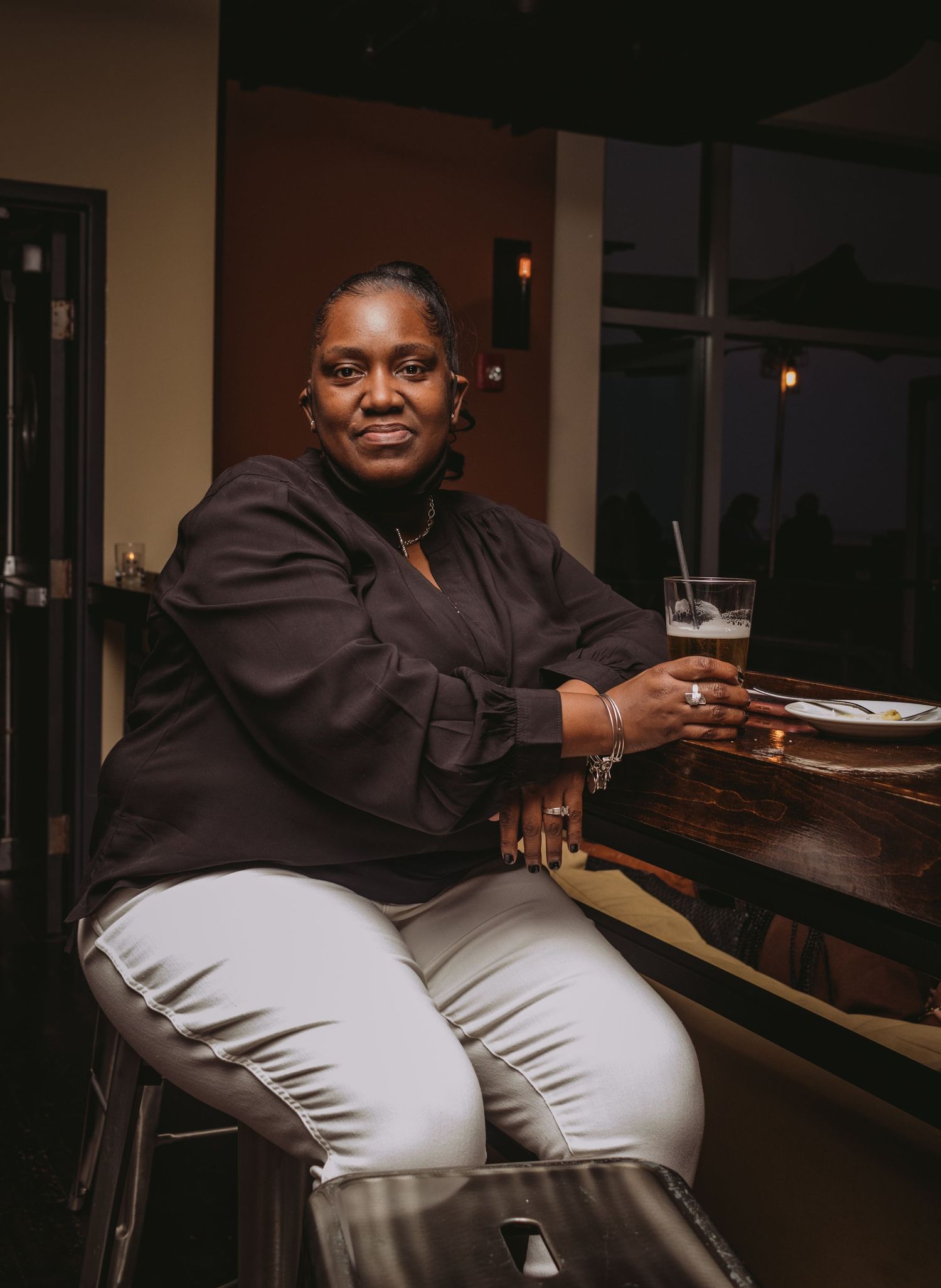 A figure wearing a dark top and light pants sits casually at a bar counter in a dimly lit setting.