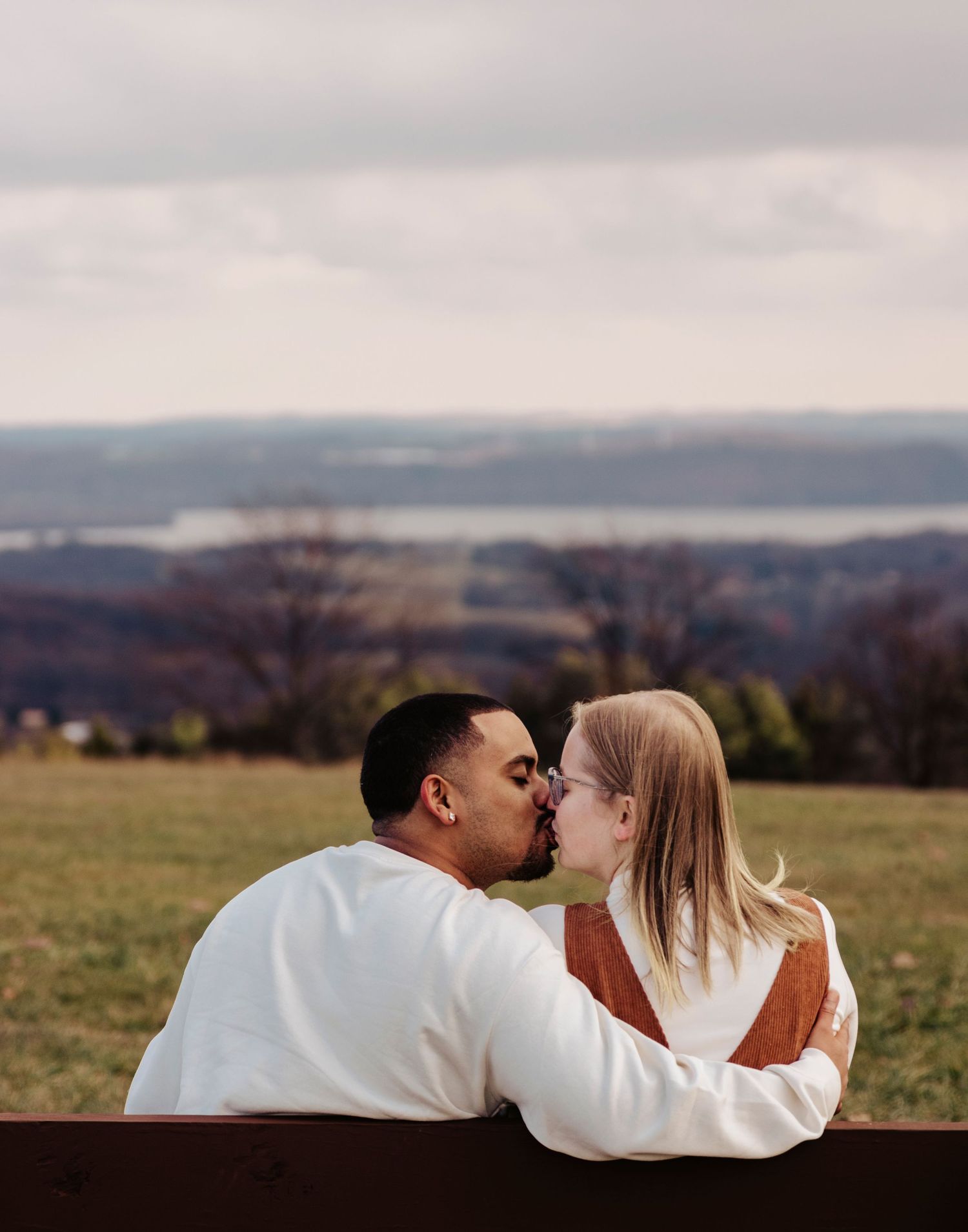 A couple shares romantic kisses in a grassy field with mountains and cloudy skies in the background.