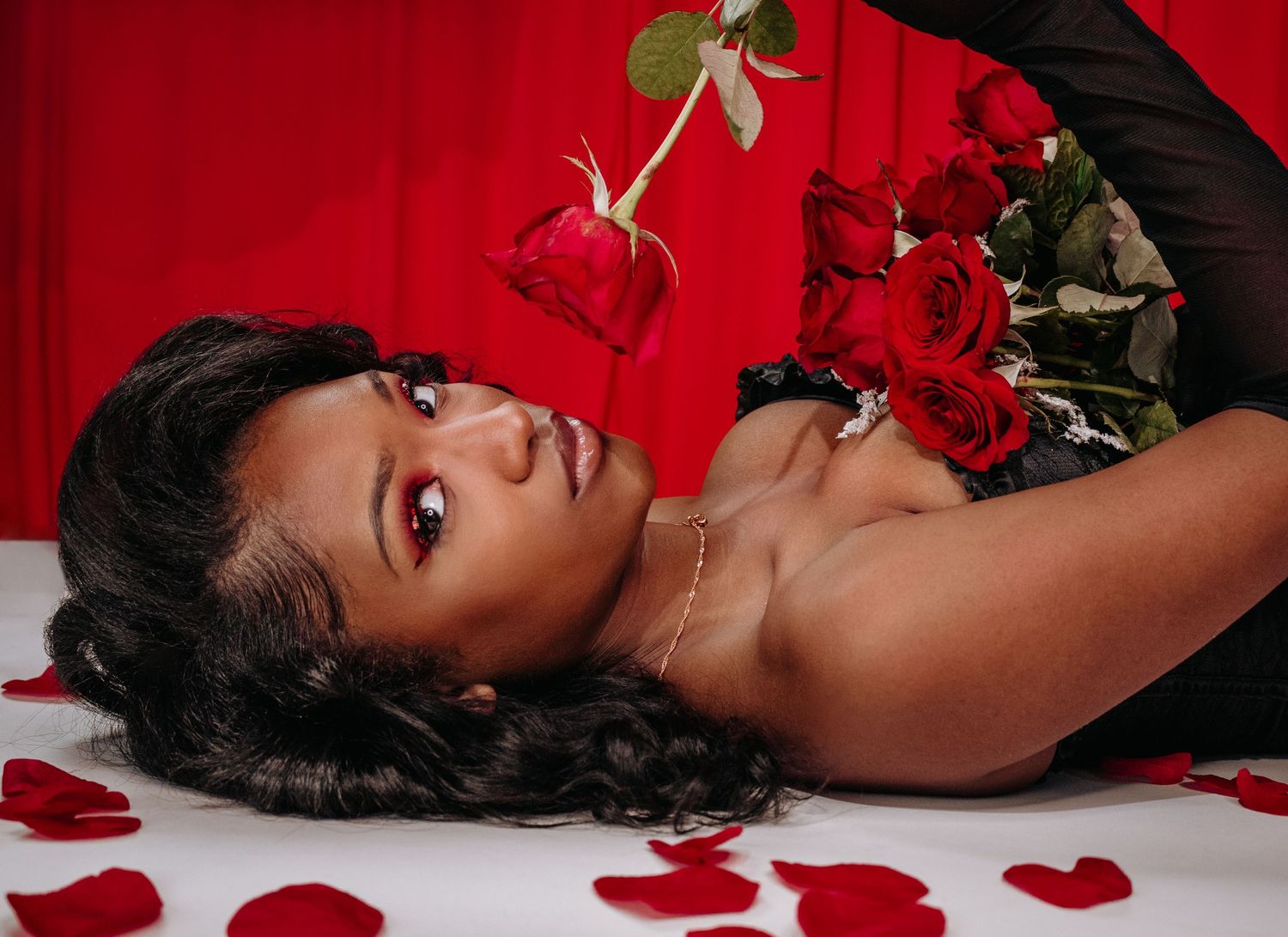 Romantic boudoir photo with red roses and petals against red backdrop.