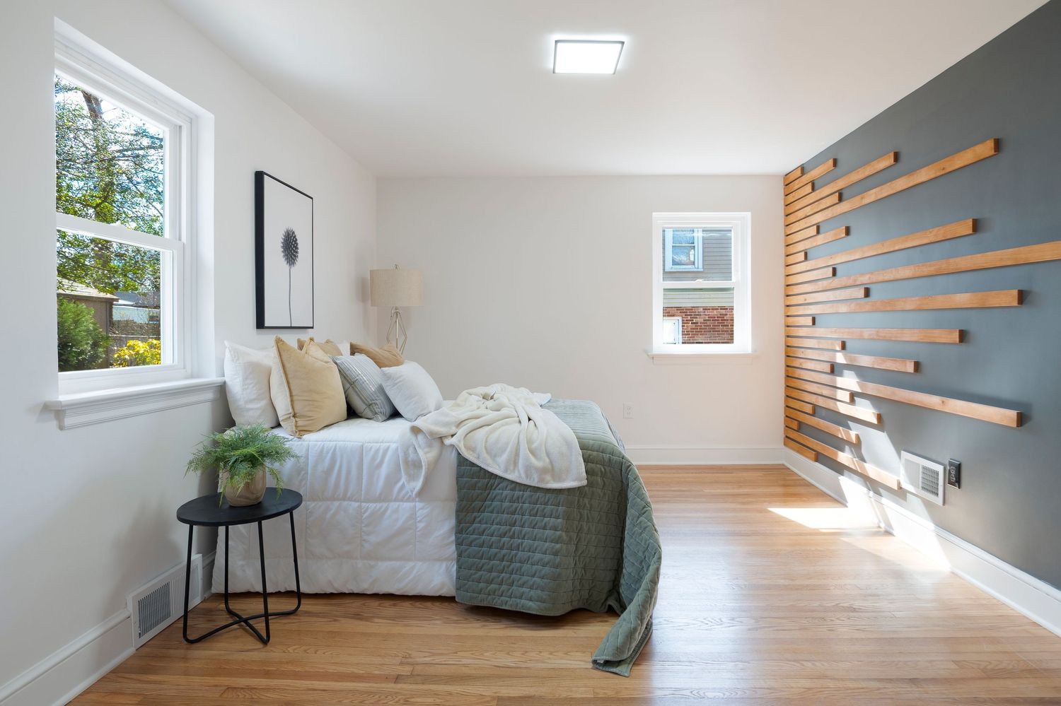 Modern bedroom with hardwood floors, white walls, wood slat accent wall, and natural light from two windows.