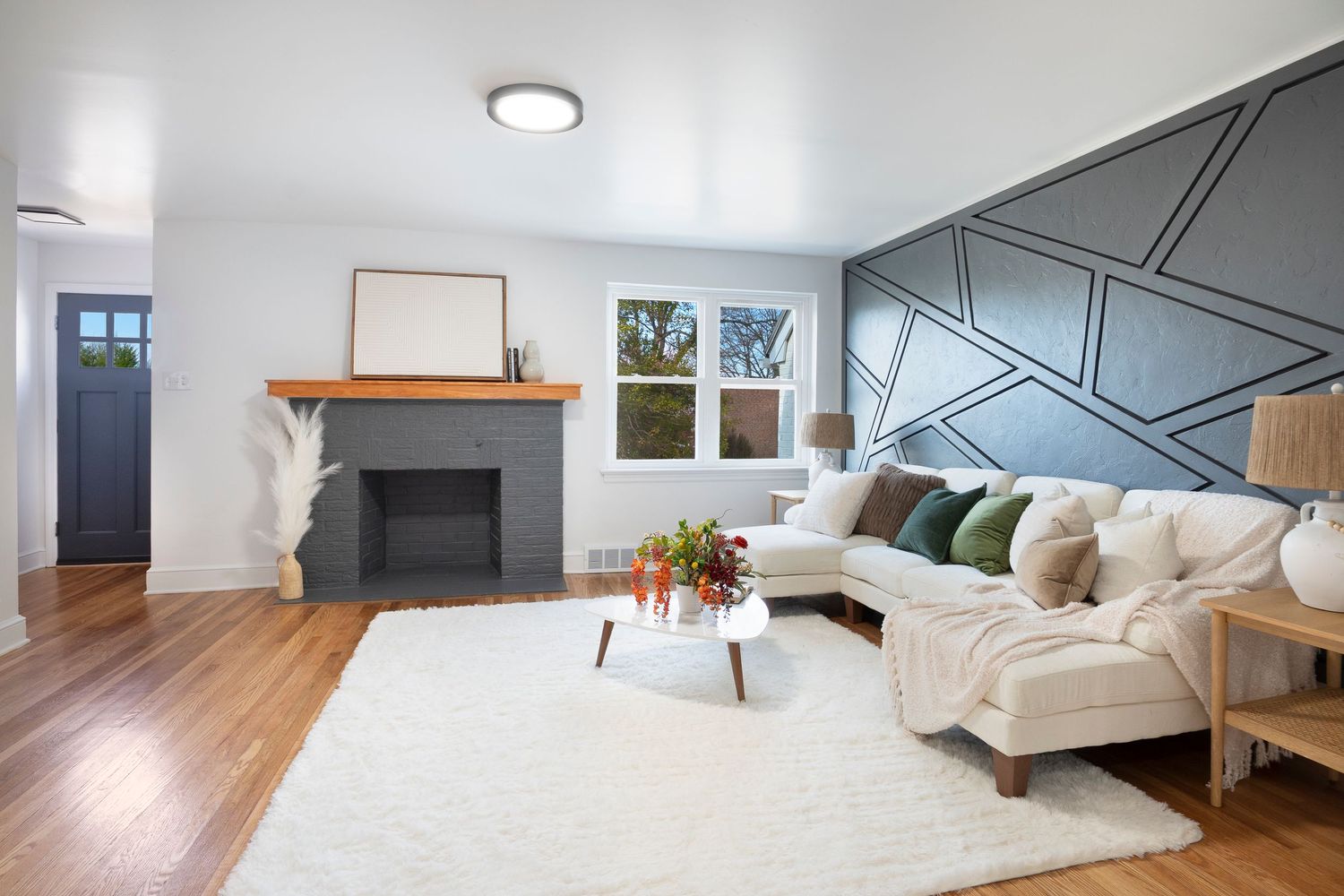 Modern living room with geometric accent wall, white sectional sofa, hardwood floors, and a gray brick fireplace with wood mantel.