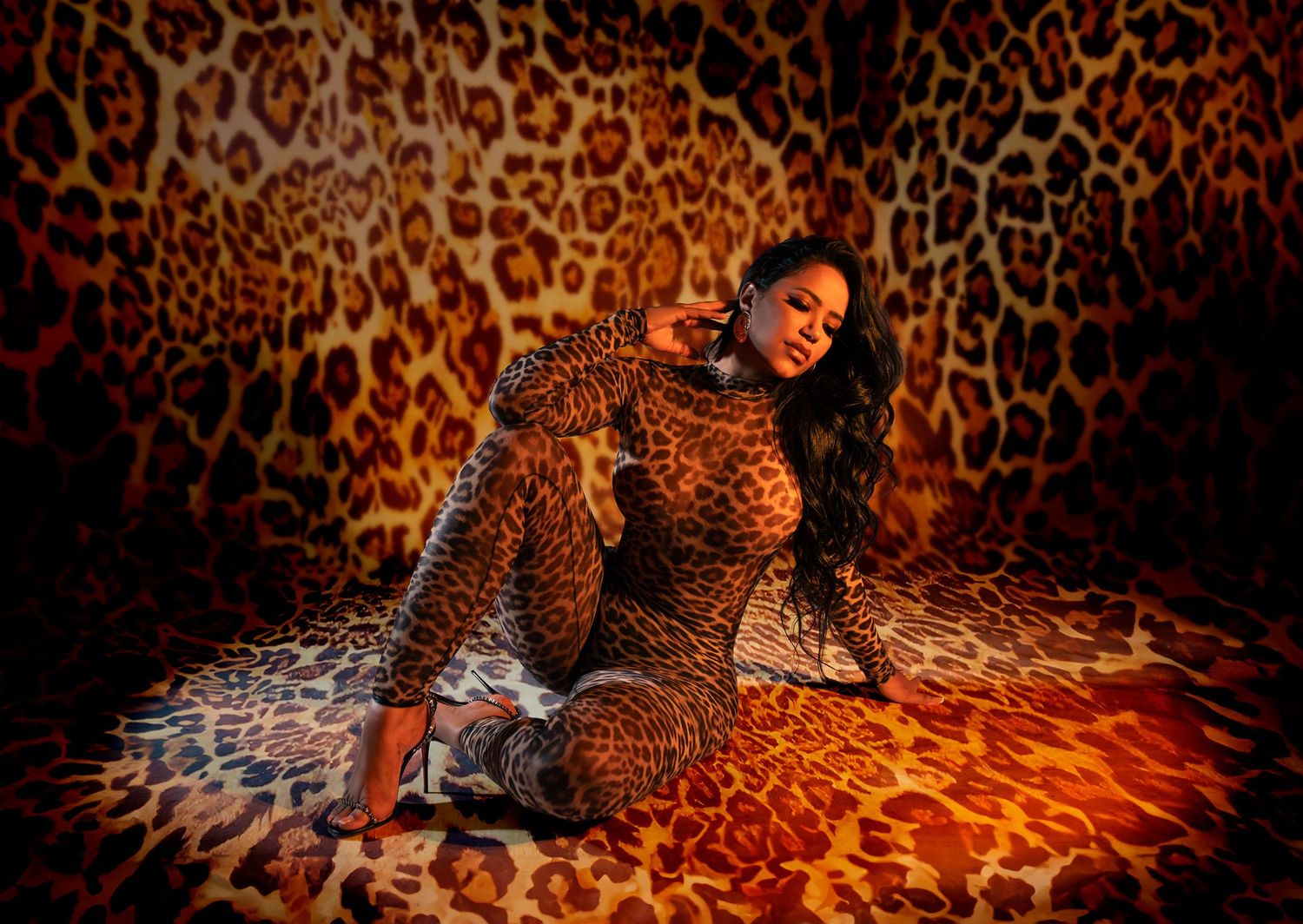 A woman in a leopard print jumpsuit dances and poses dynamically in front of a matching leopard print background.