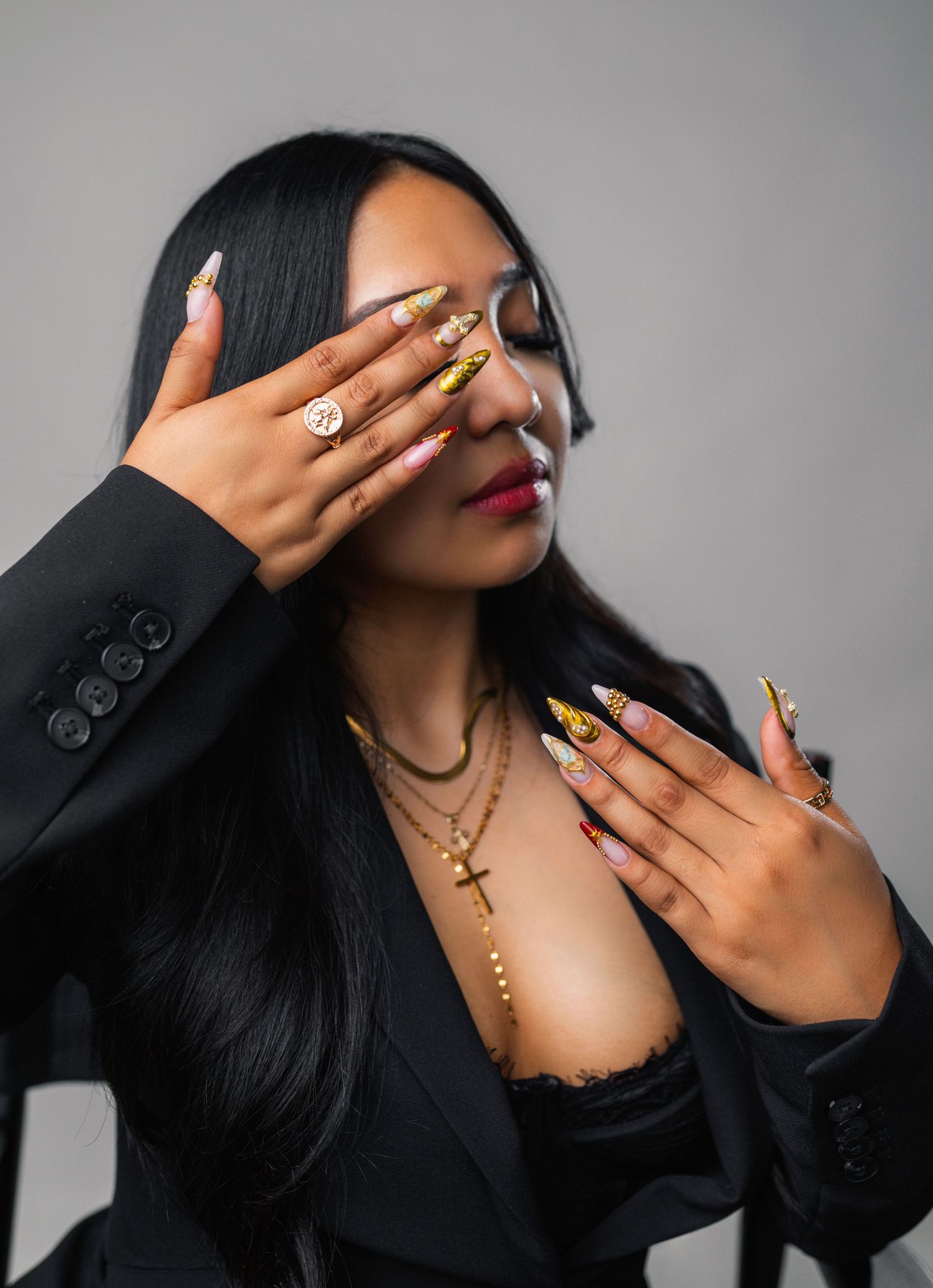 A model wearing black poses with gold nails and jewelry against a gray backdrop in artistic poses.