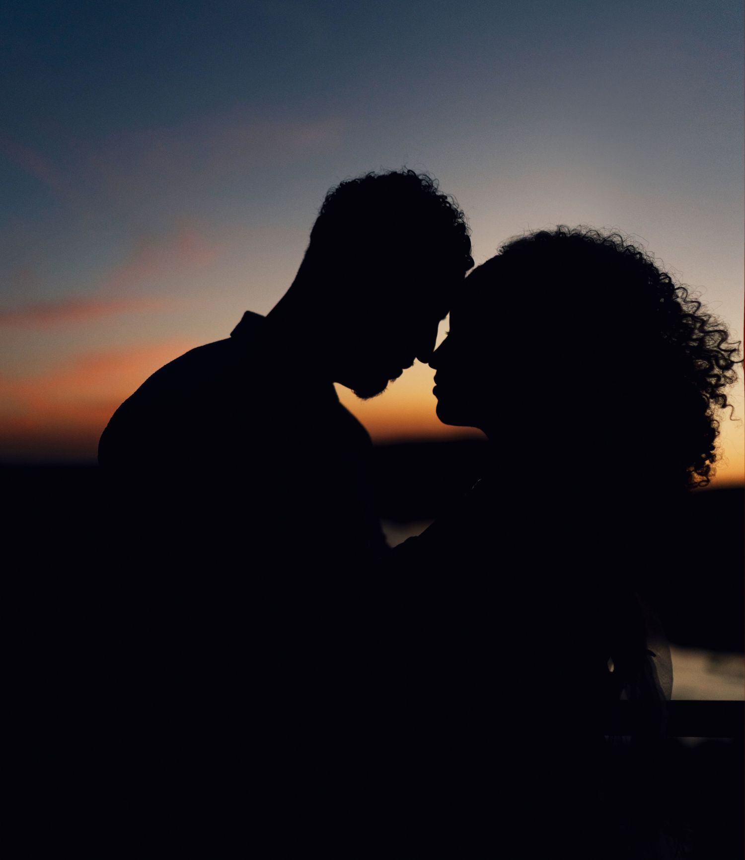 Silhouetted figures in a romantic embrace against a dramatic sunset sky over water.