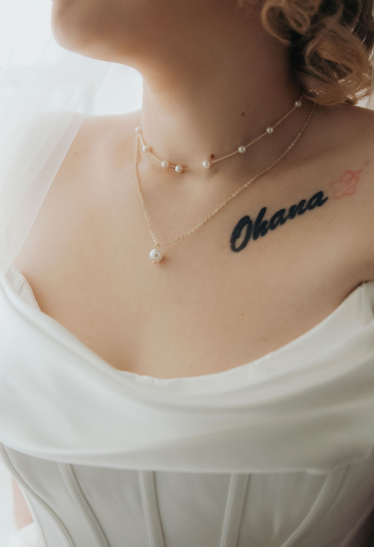 Close-up of bride wearing pearl necklaces and an Ohana chest tattoo in a white wedding dress.