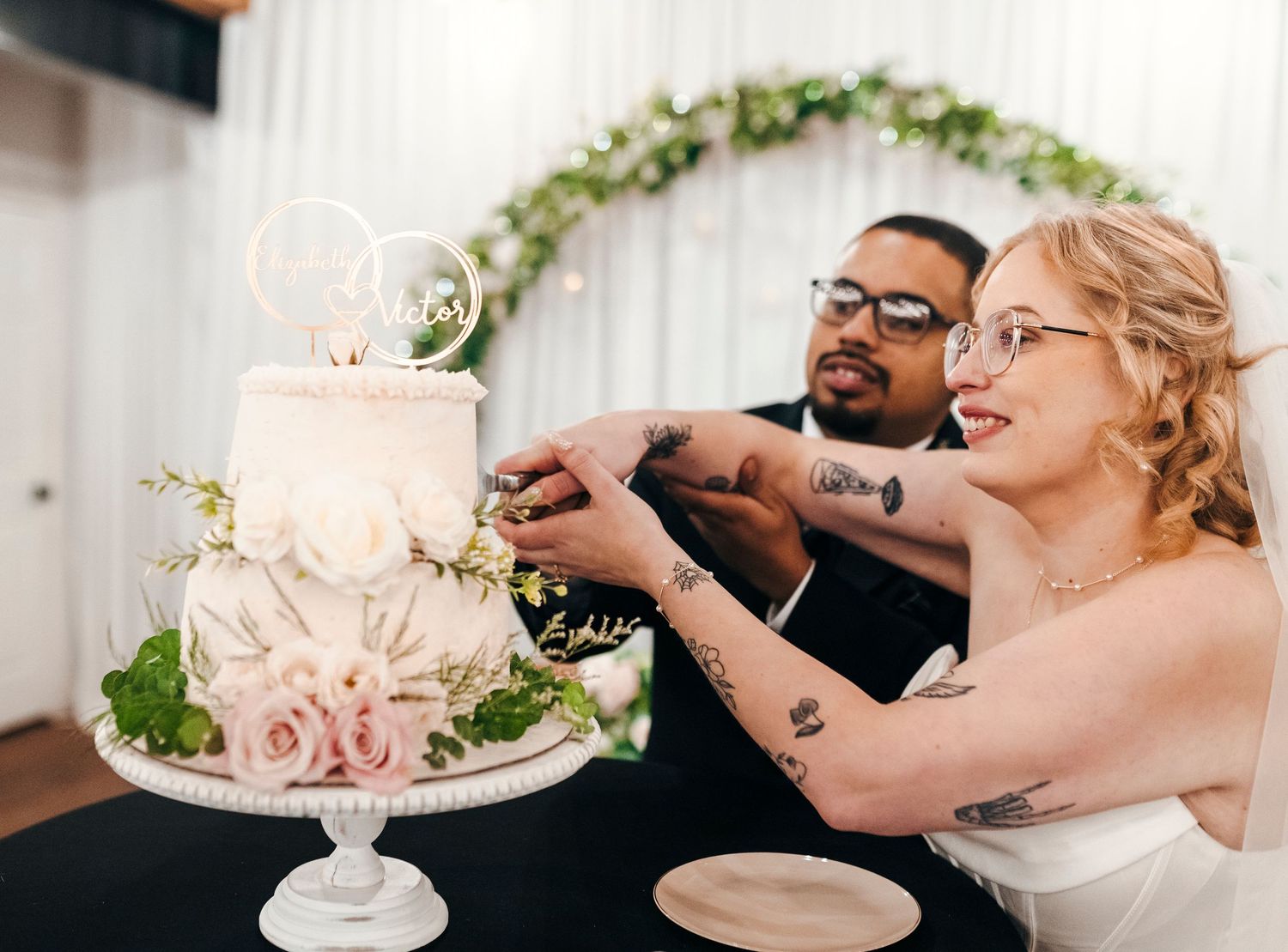 Bride and groom cutting elegant two-tier white wedding cake decorated with pink roses and greenery.