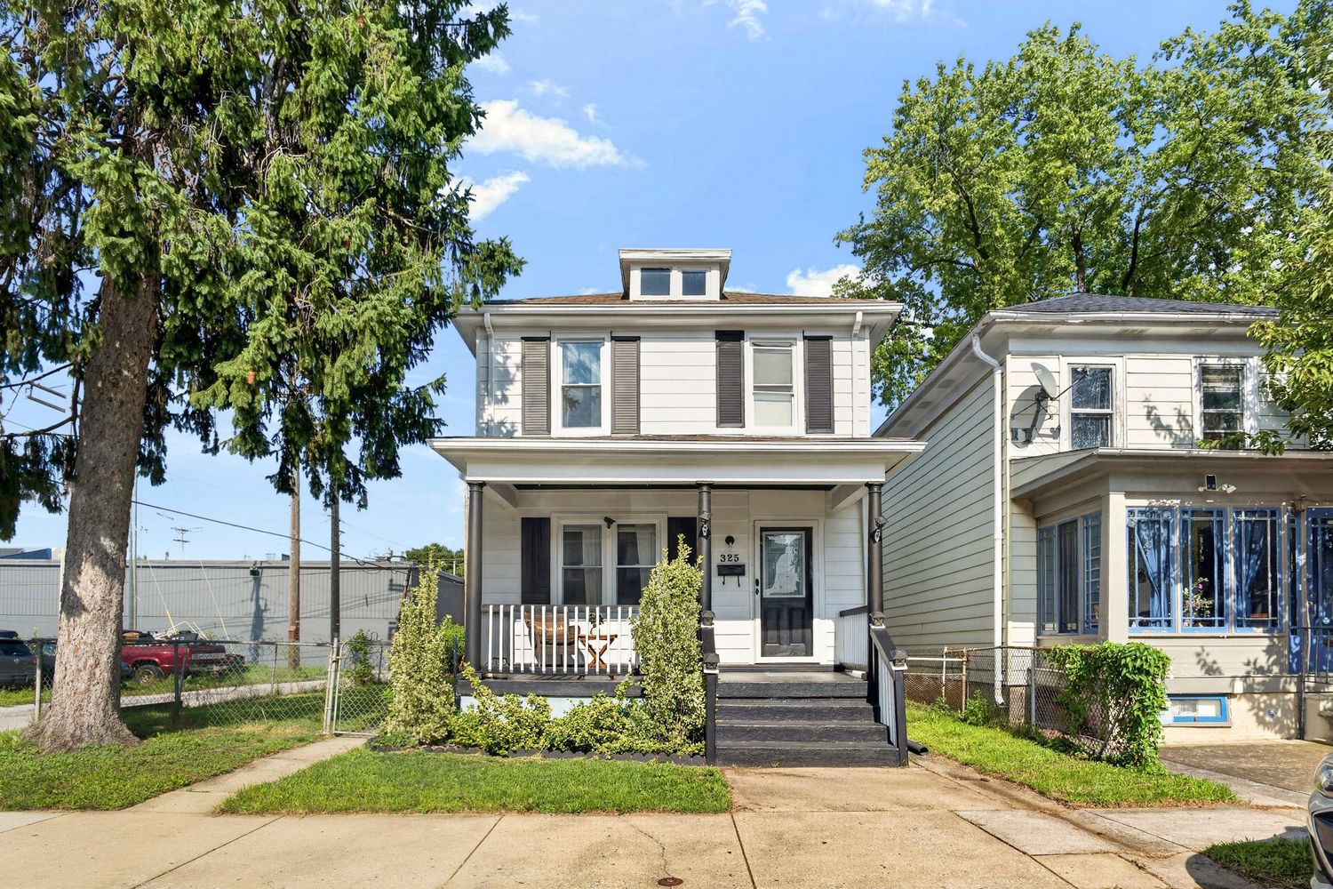 Two-story gray American Foursquare home with white porch railings, mature trees, and manicured lawn on a sunny day.