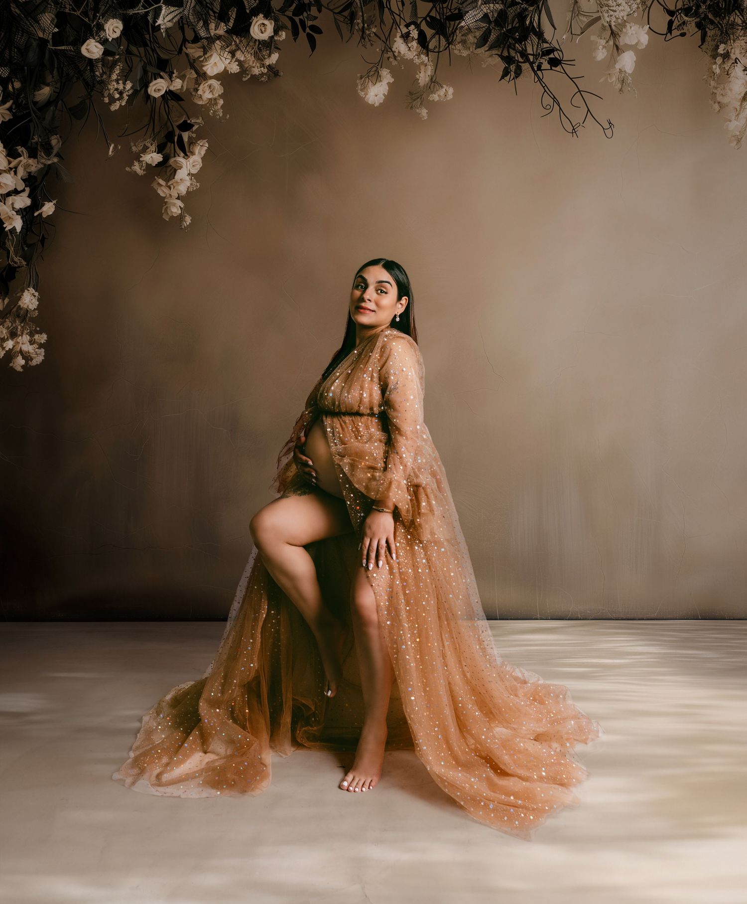 A glamorous maternity photo featuring flowing peach-colored lace gown against a moody backdrop with floral accents.