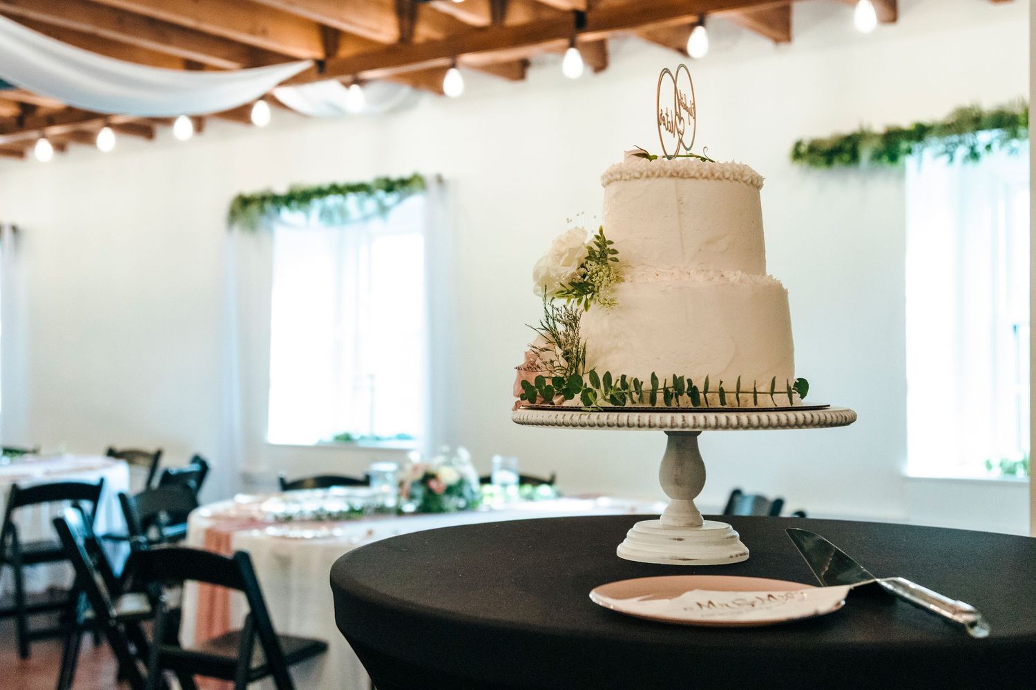 Elegant two-tier white wedding cake with floral decor on a pedestal stand in a decorated reception hall.