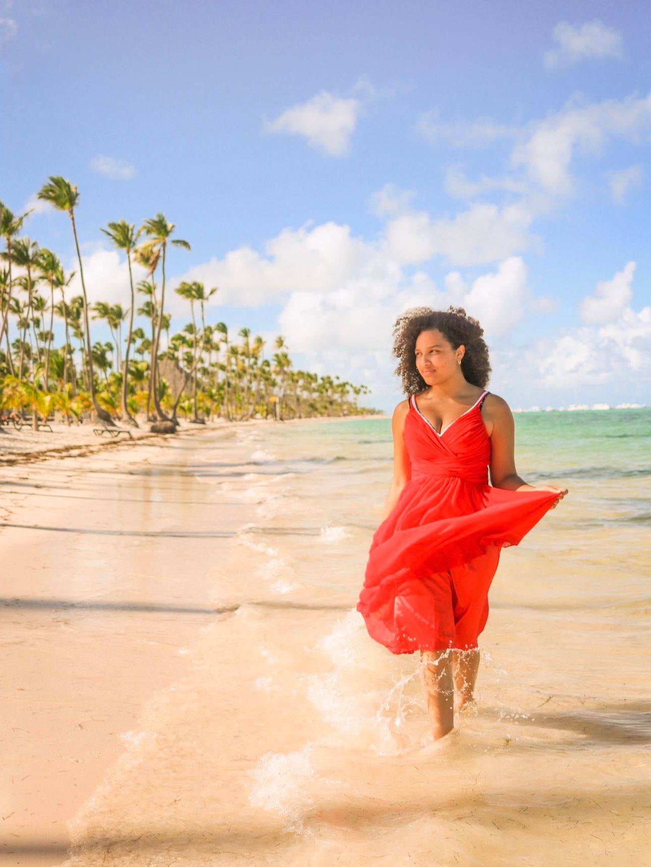 Woman in flowing red dress runs joyfully along tropical beach lined with palm trees.