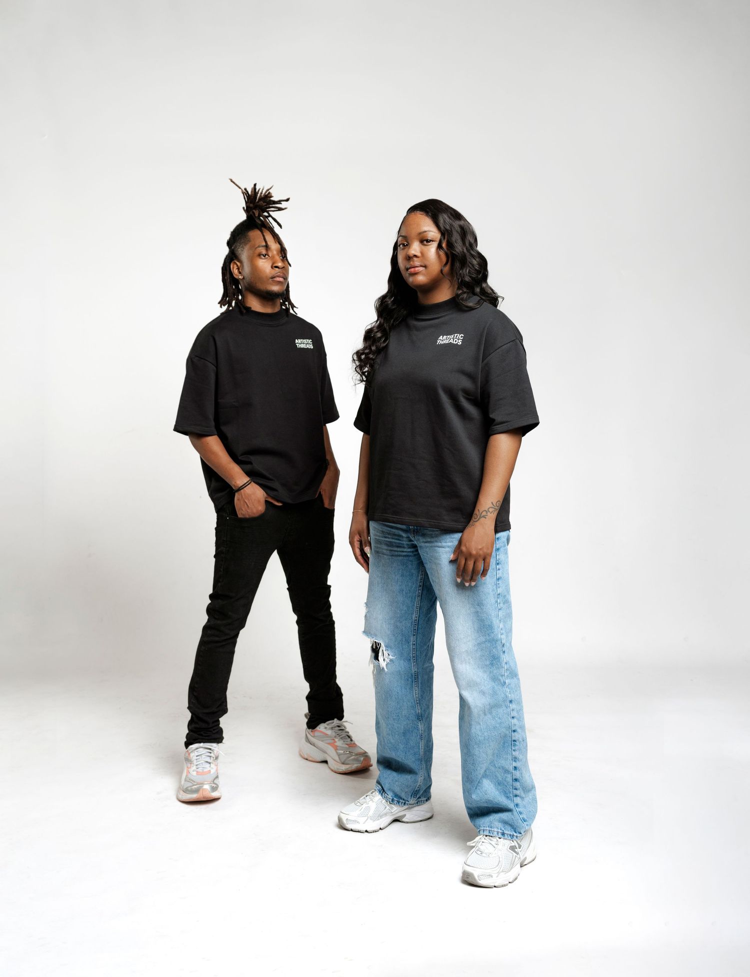 Two people in black t-shirts and jeans stand together in a stylish pose against white background.