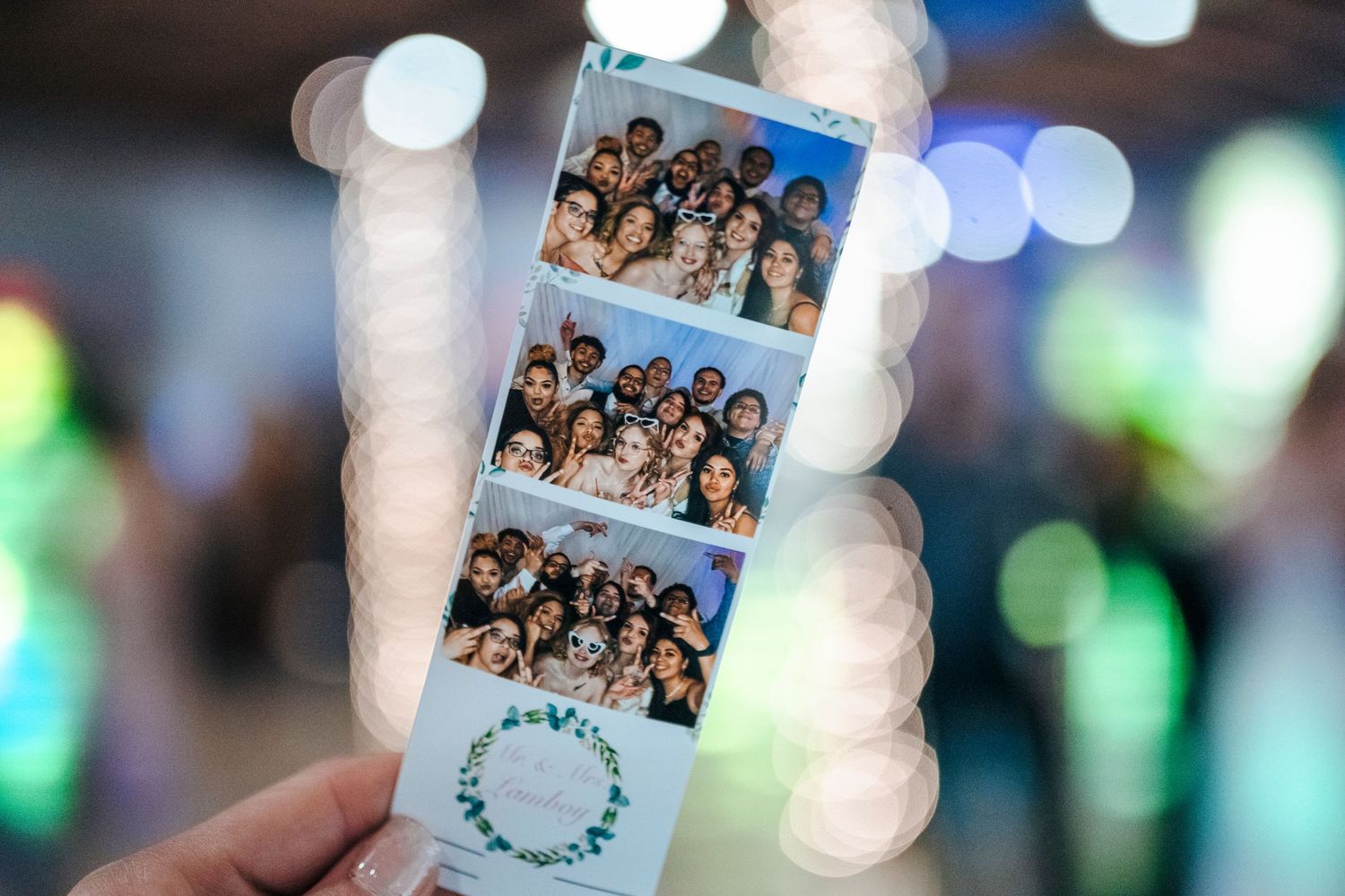 Hand holding a photo booth strip with group photos from a wedding reception with bokeh background.