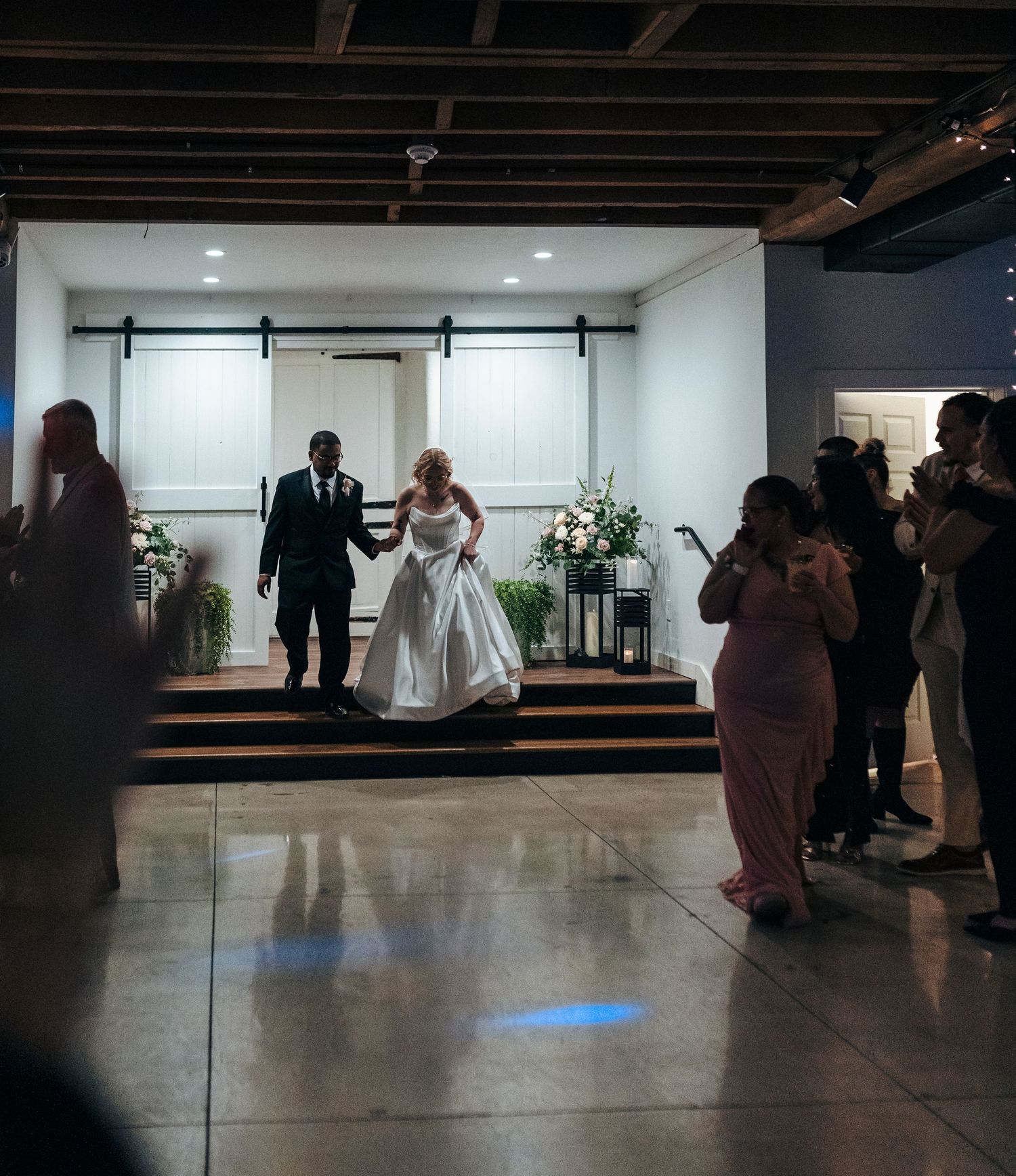Bride and groom make grand entrance into dimly lit wedding reception venue while guests cheer and photograph.