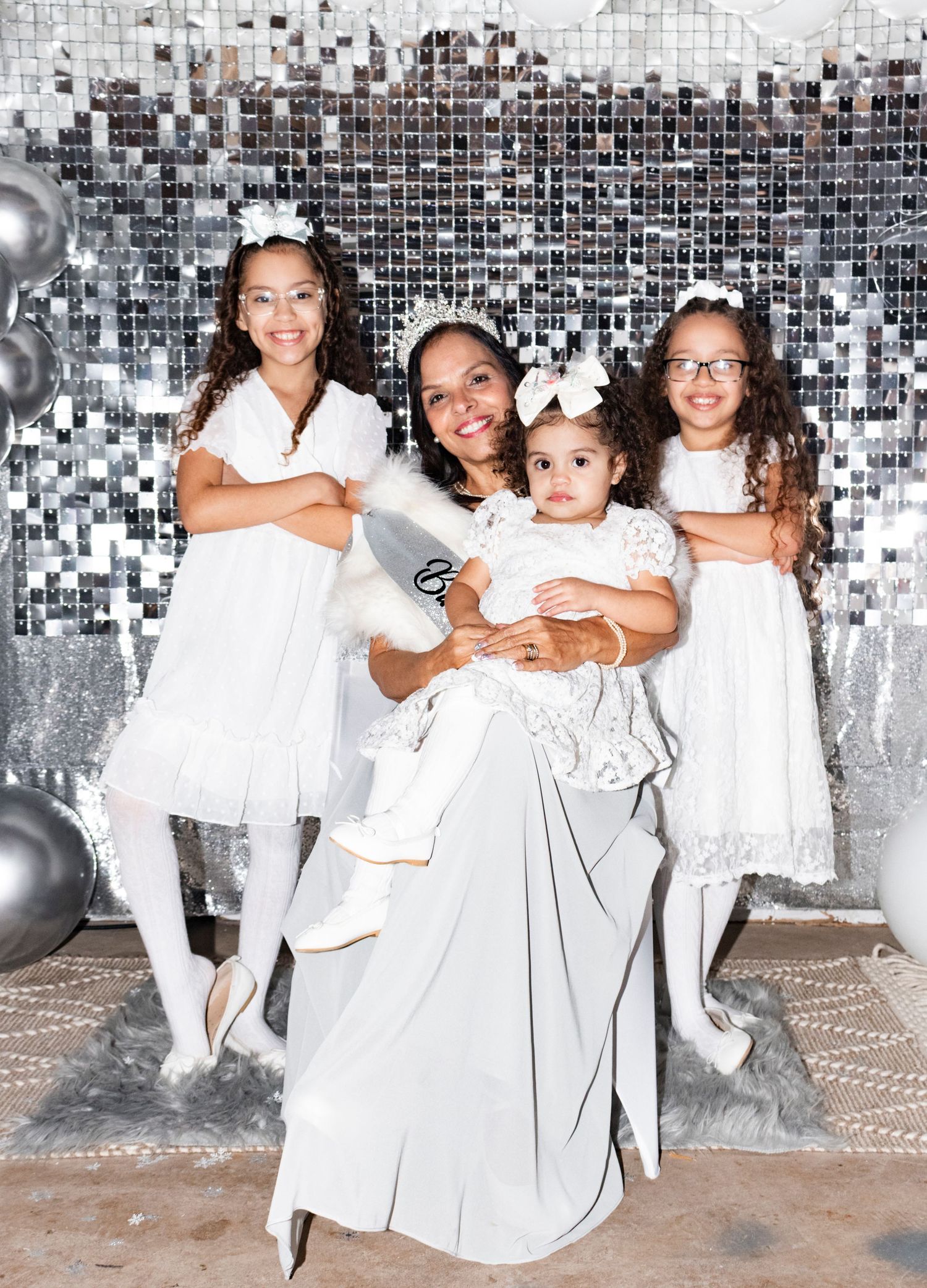 A group in sparkling white dresses poses together against a silver sequined backdrop with gray balloons.