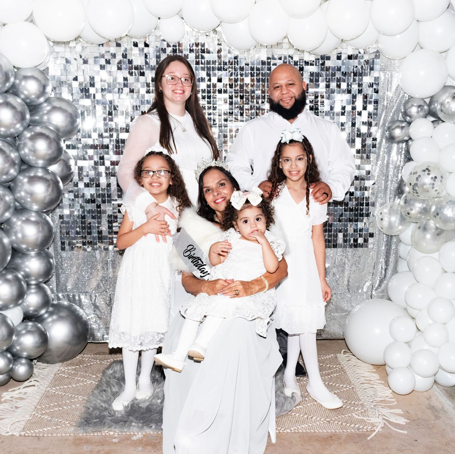 Family in coordinated white outfits pose against a festive silver and white balloon backdrop for holiday photos.