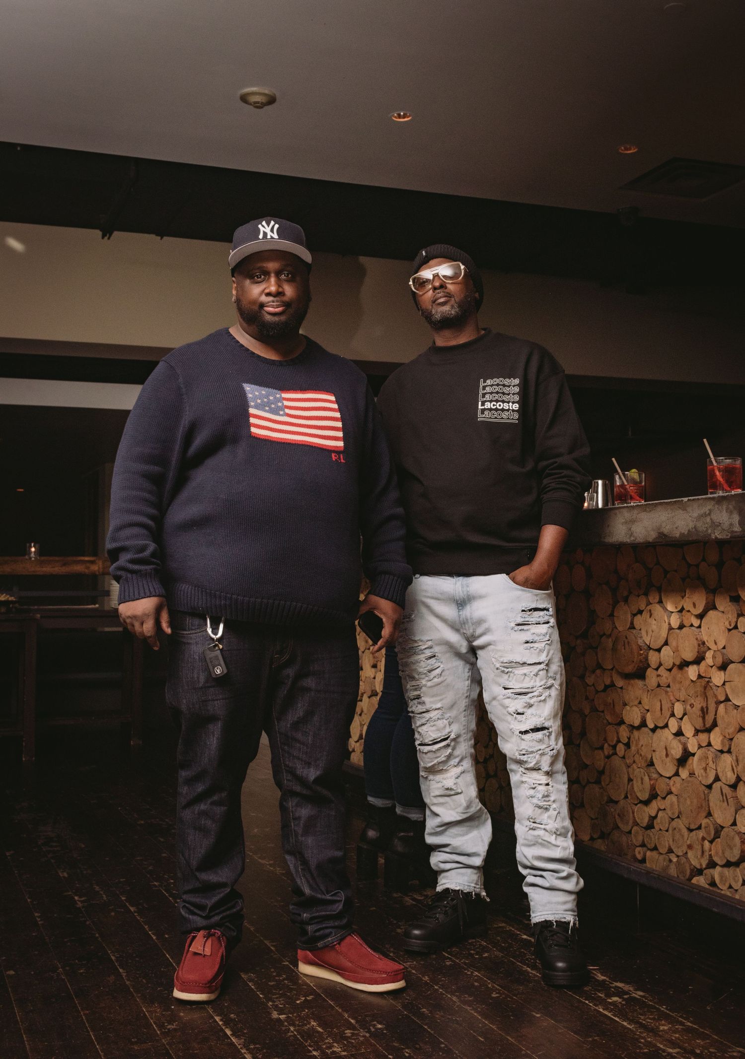 Two people in contrasting outfits of navy blue and light gray stand against a rustic stone wall background in dim lighting.