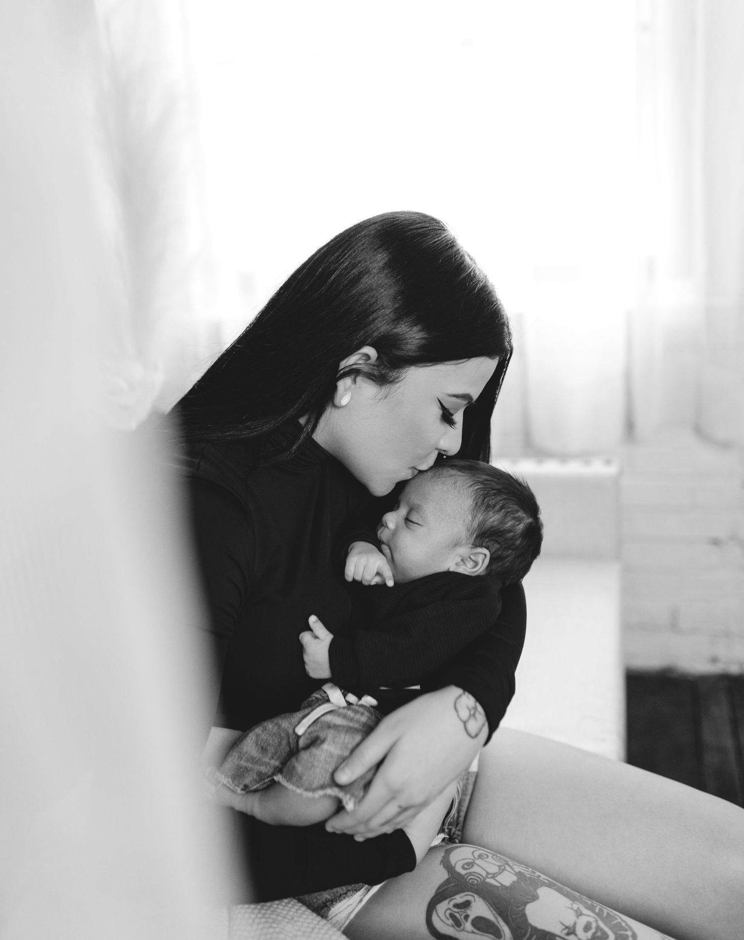 Black and white photo of a dark-haired mother tenderly kissing her newborn baby while cradling it in her arms.