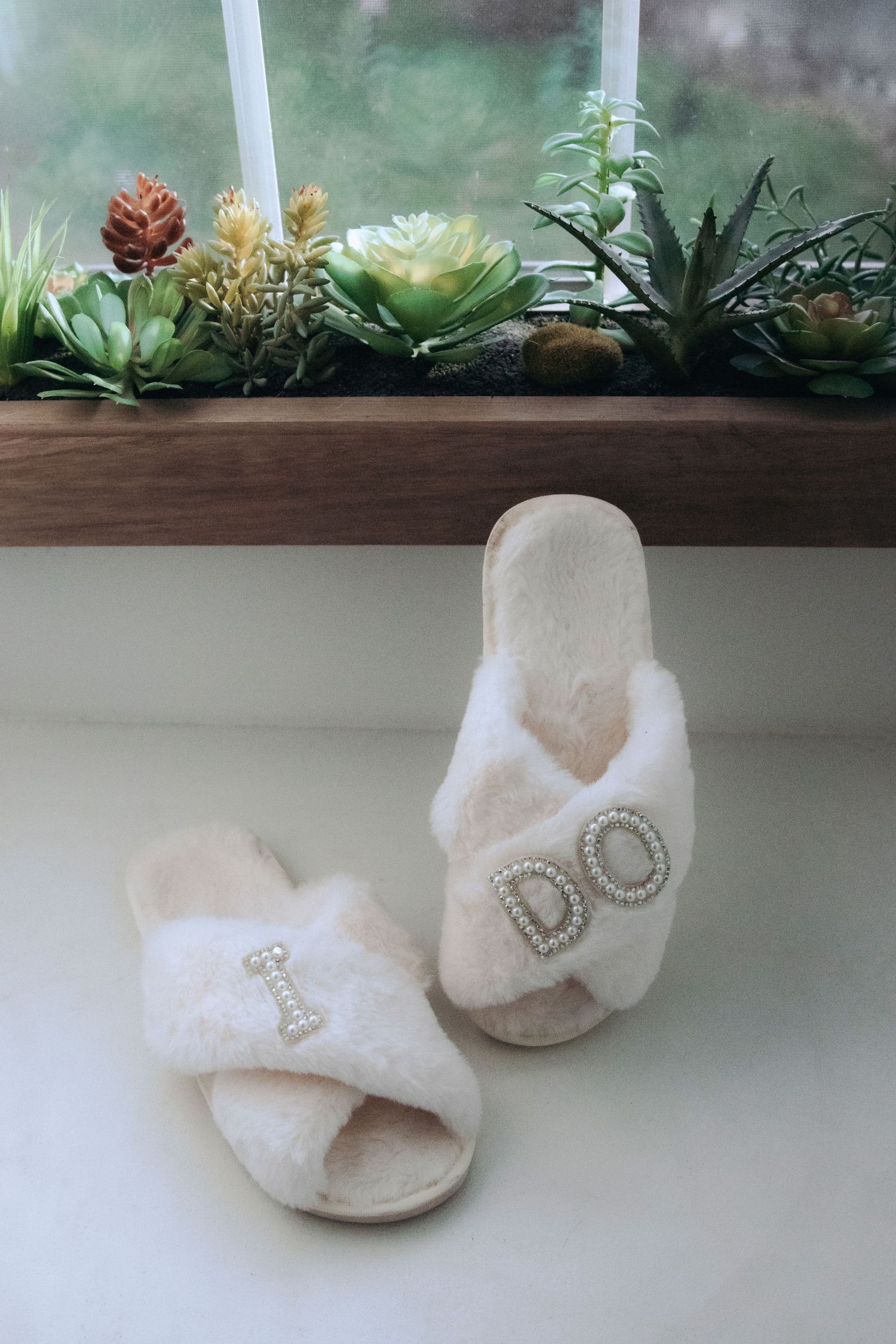 White fluffy bridal slippers with pearl letter embellishments spelling I DO, displayed on windowsill with succulents.