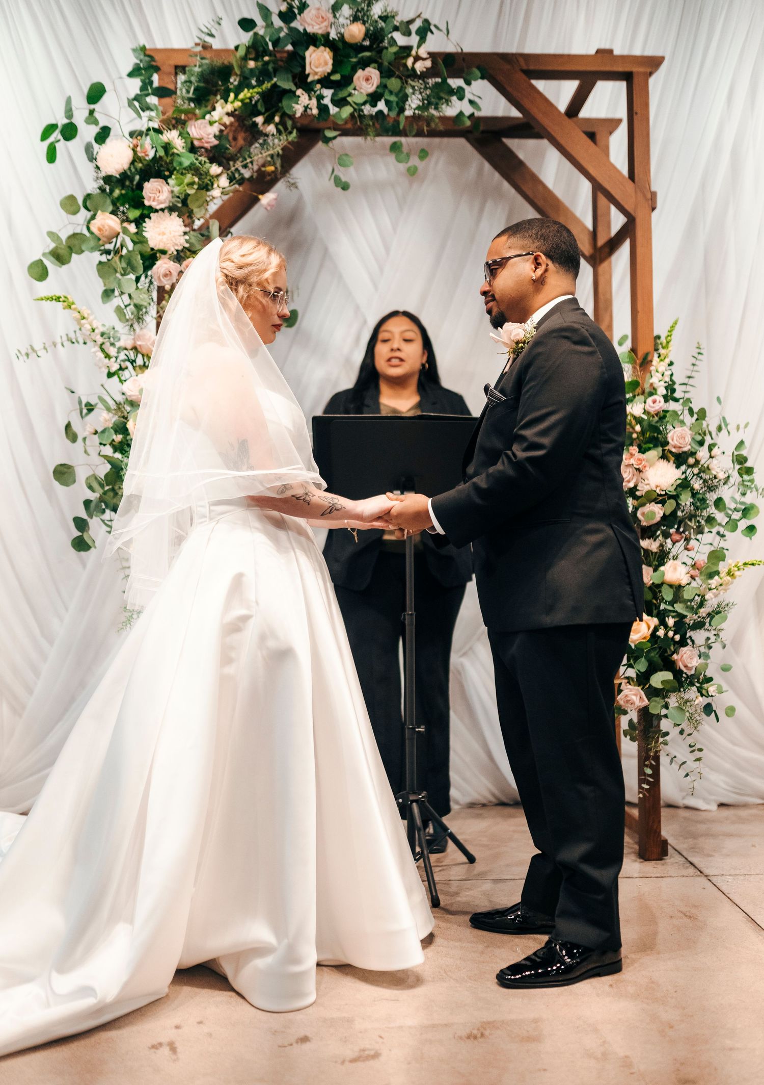 Bride and groom exchange vows under a wooden arch with blush floral arrangements at wedding ceremony.