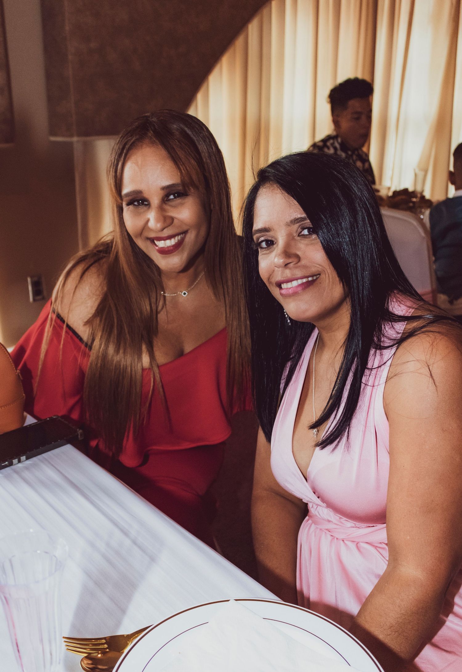 Two women in red and pink dresses smiling while seated at an elegant dinner table.