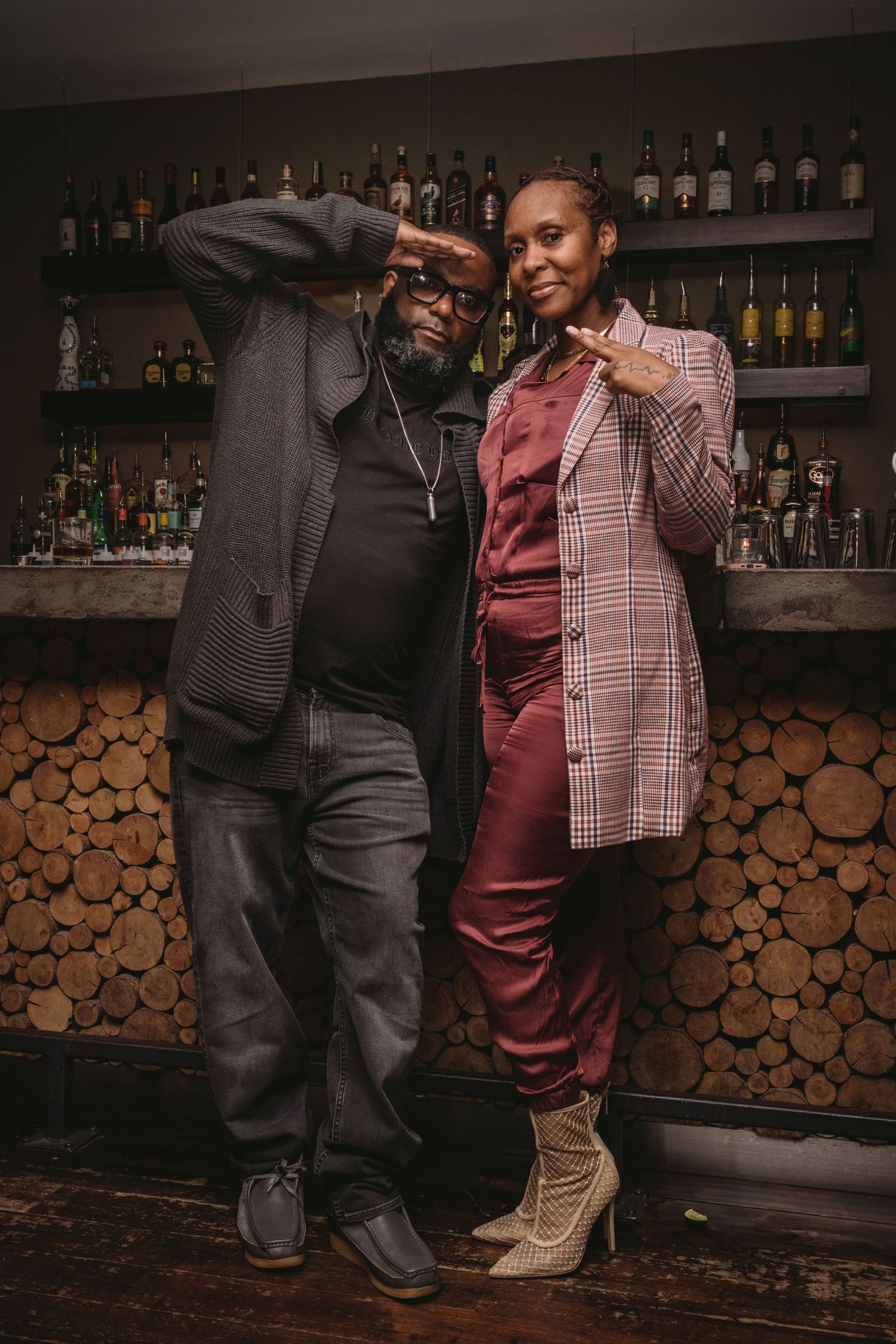 Two people pose together in stylish outfits at a bar with wooden decor and bottles displayed on shelves behind them.