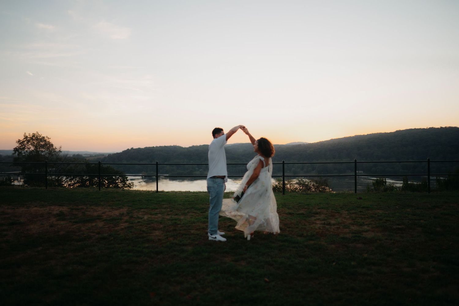 Couple in white attire walks through scenic mountain landscape during golden hour sunset.