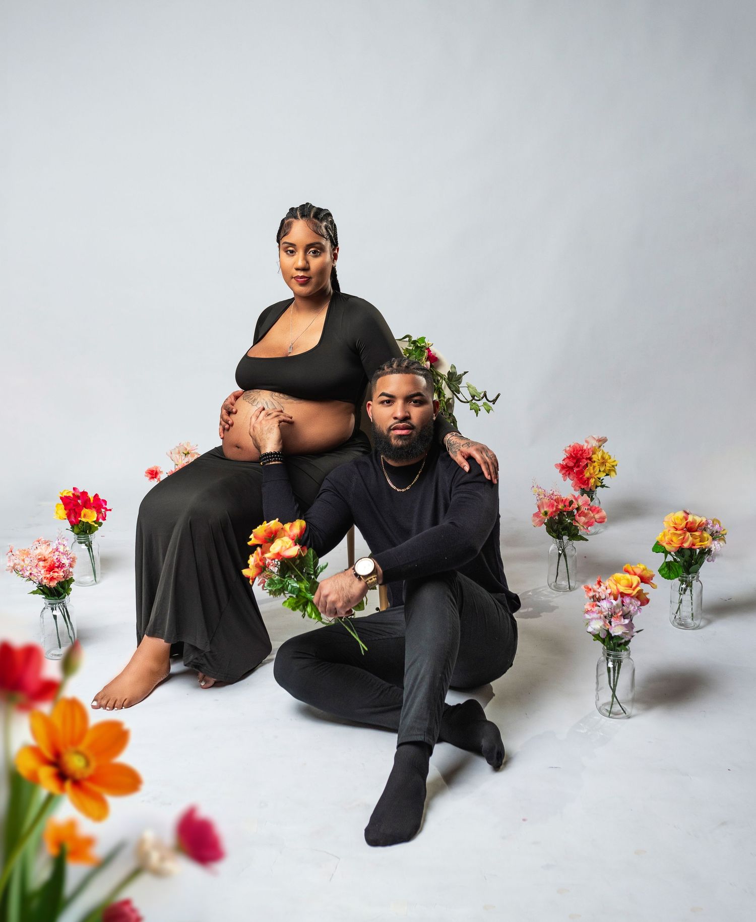 Maternity photoshoot featuring couple in black attire surrounded by colorful flowers in glass vases against light backdrop.
