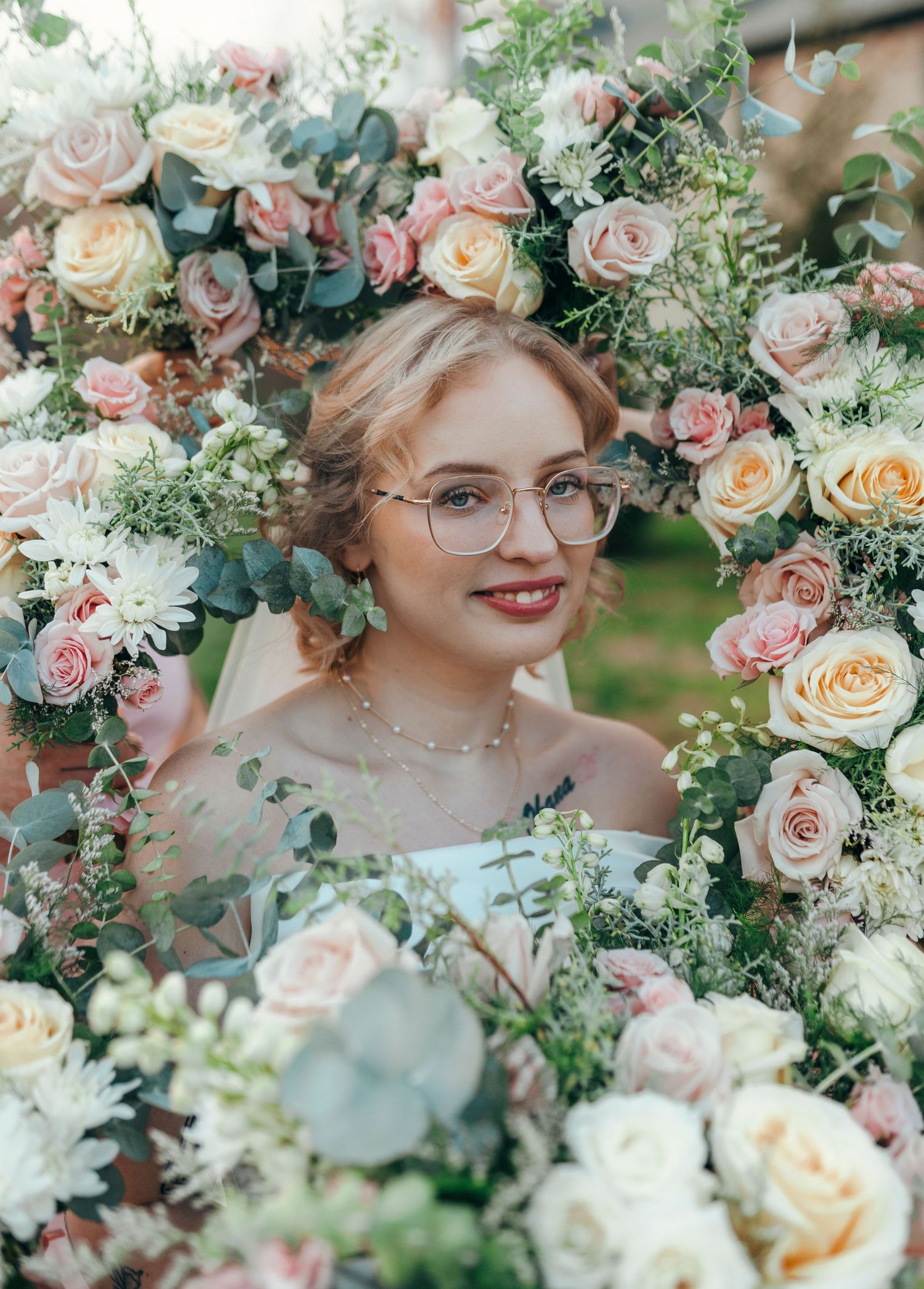 Smiling bride with glasses surrounded by lush pink, white, and peach rose bouquets on her wedding day.
