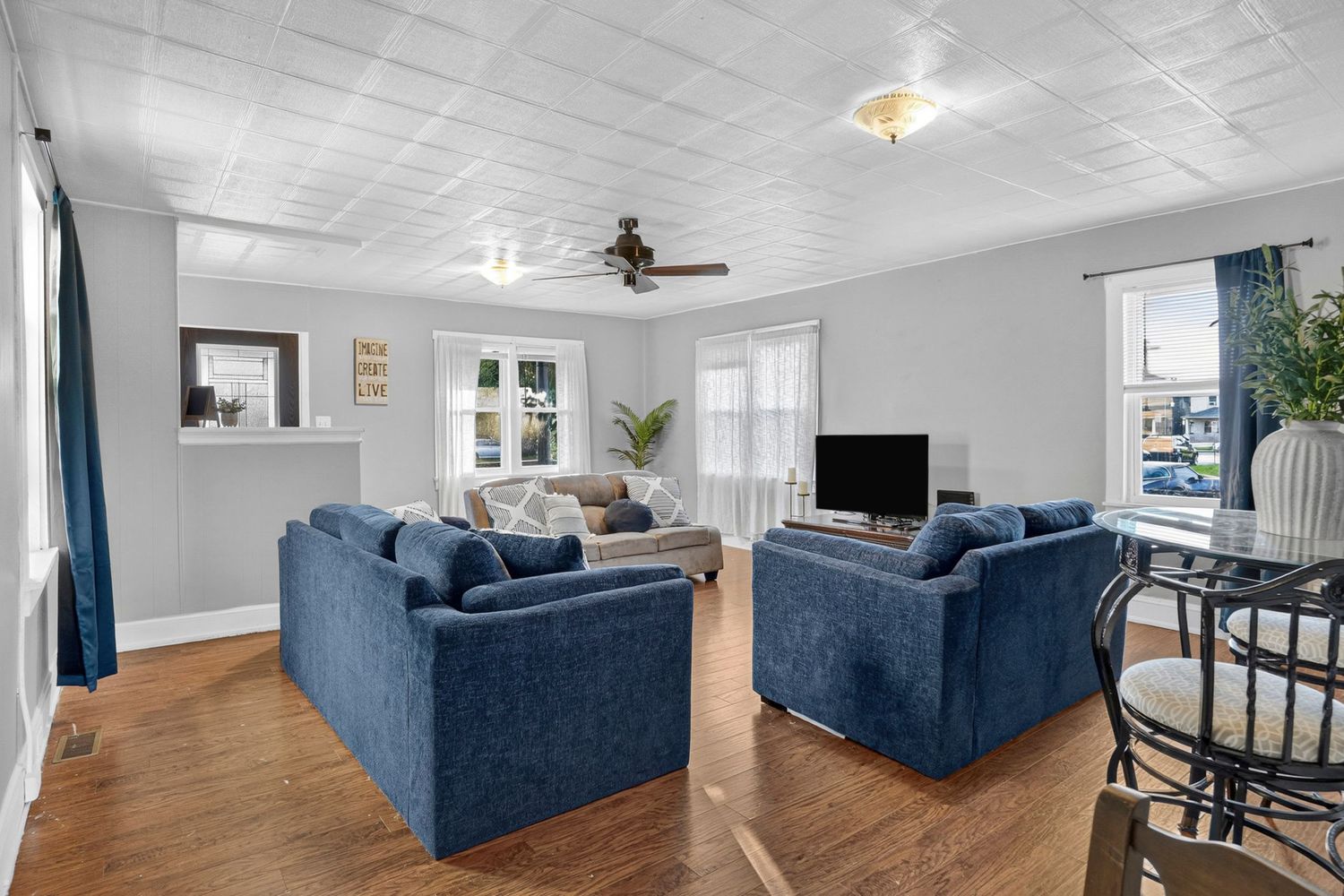 Bright living room with blue sofas, hardwood floors, ceiling fan, and large windows filling the space with natural light.