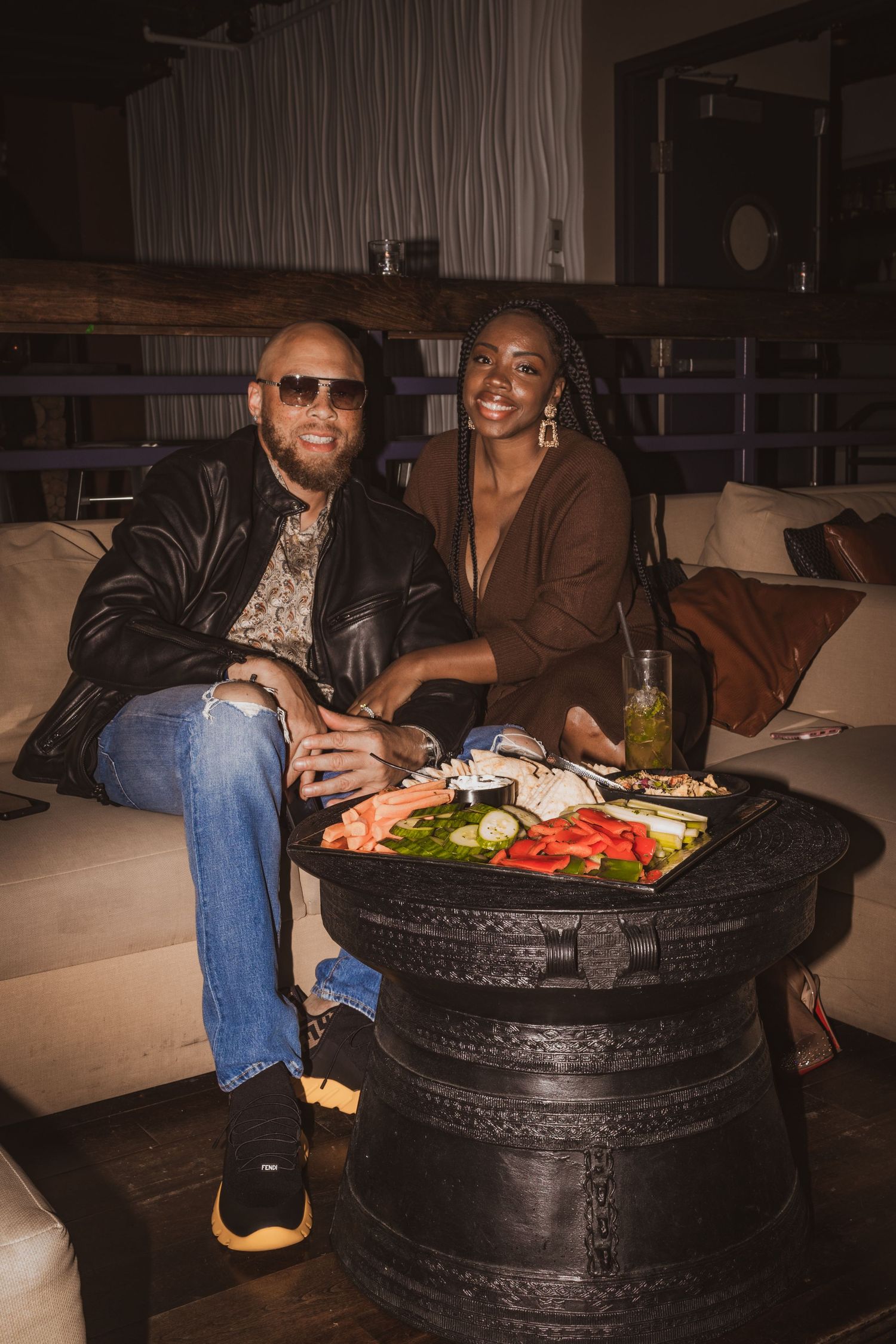 People enjoying appetizers and drinks on a couch with a unique tire-made coffee table in a dimly lit room.