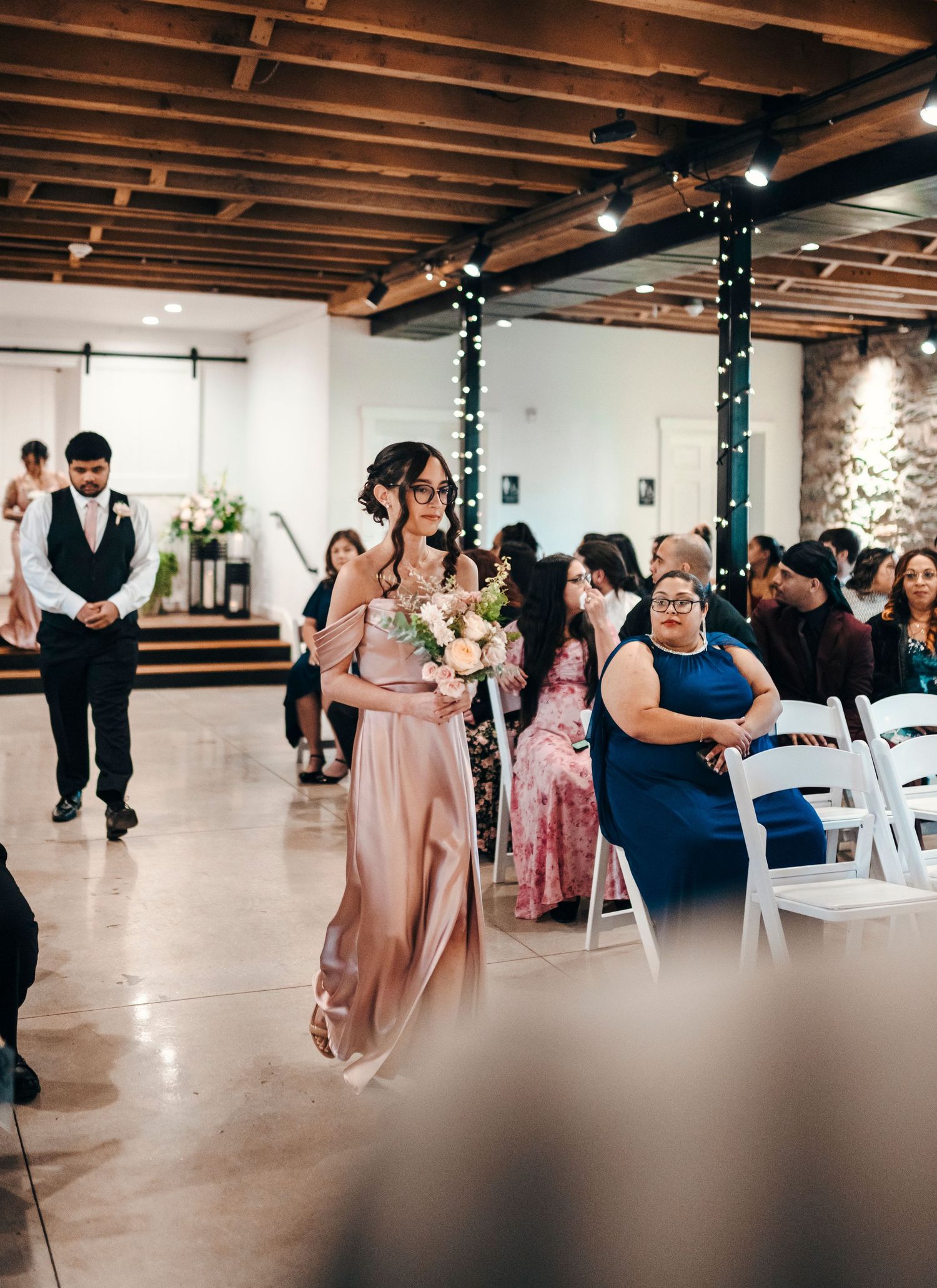Bridesmaid in blush pink gown walks down aisle holding bouquet at elegant indoor wedding ceremony.