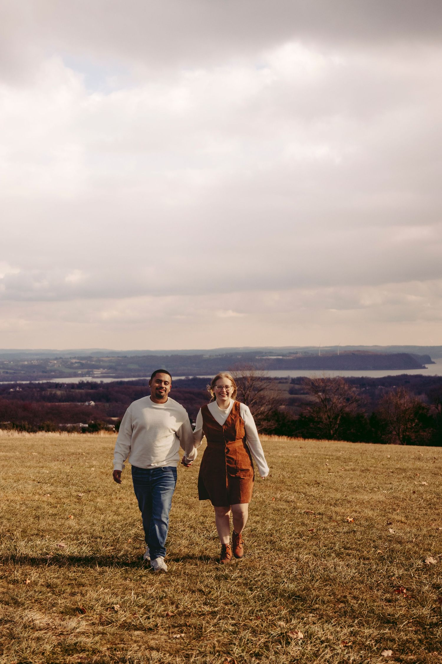 Two people hold hands while walking together across a golden autumn field with rolling hills in the background.