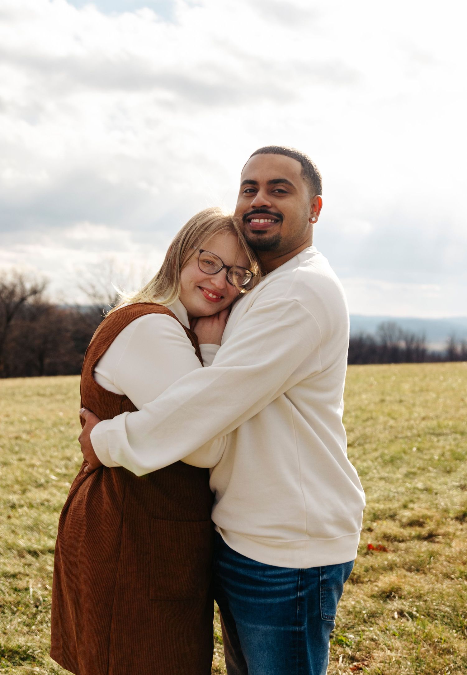 A couple embraces while standing in a grassy field on an overcast day, with mountains visible in the background.