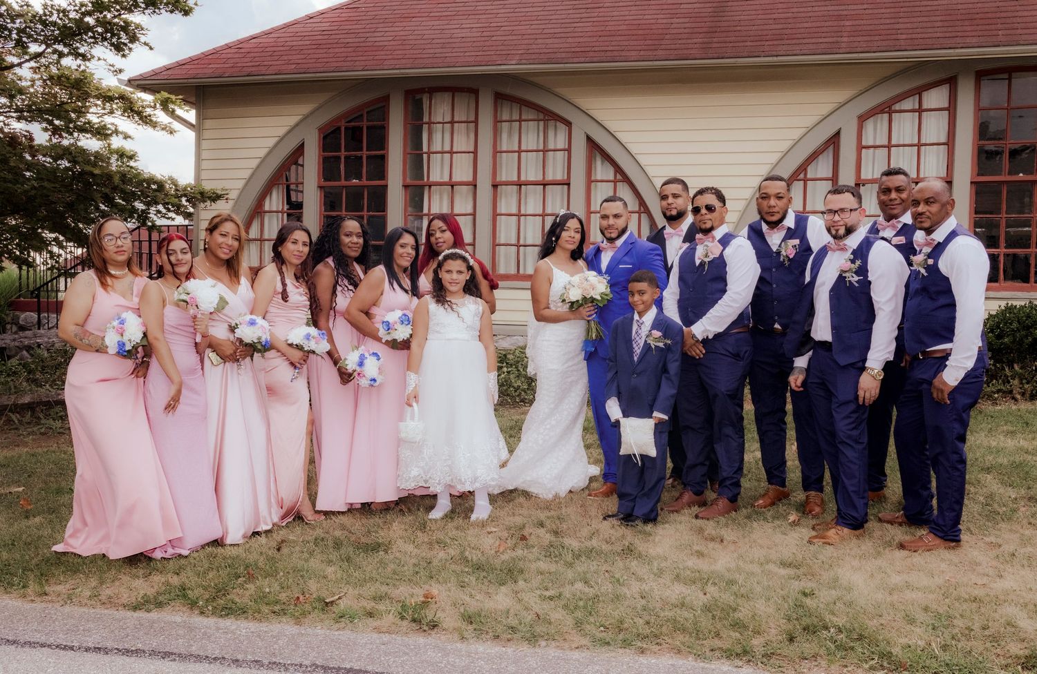 Large wedding party in pink dresses and blue vests posing by arched windows.